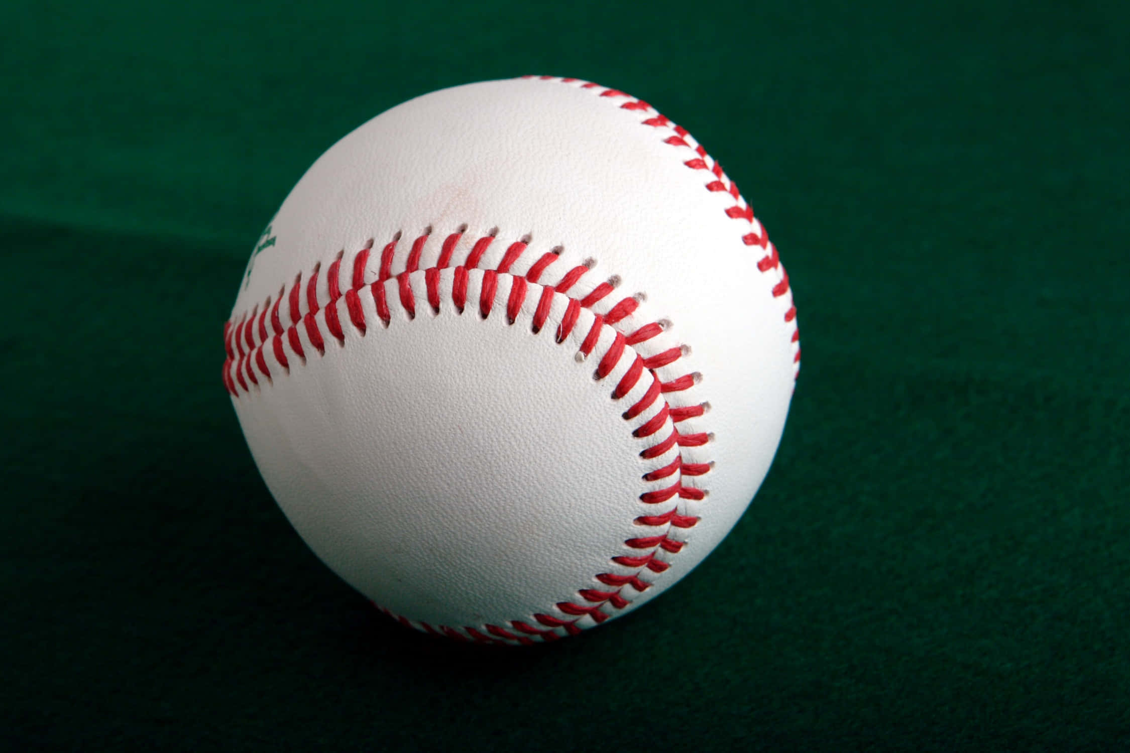 Cool Baseball 桌布 壁紙 — 一個帶有紅色縫線的純白色棒球，在柔和燈光下放置在深綠色絨氈表面上。