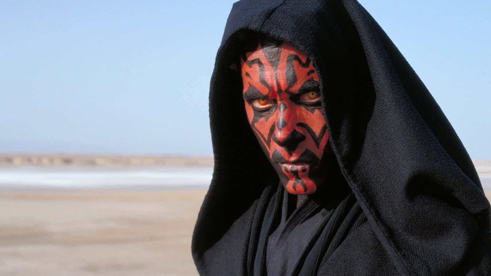 The Phantom Menace 桌布 壁紙 — 一個帶有紅色臉部彩繪和發光黃色眼睛的威脅性人物，站在晴朗藍天下的一片貧瘠沙漠中。