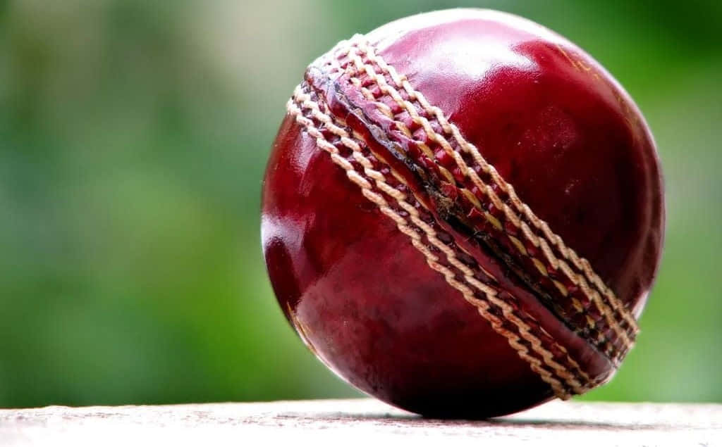 Cricket 桌布 壁紙 — 一個帶有縫線的光澤紅色皮革板球近距離特寫，放置在木質表面上，背景為模糊的綠色。
