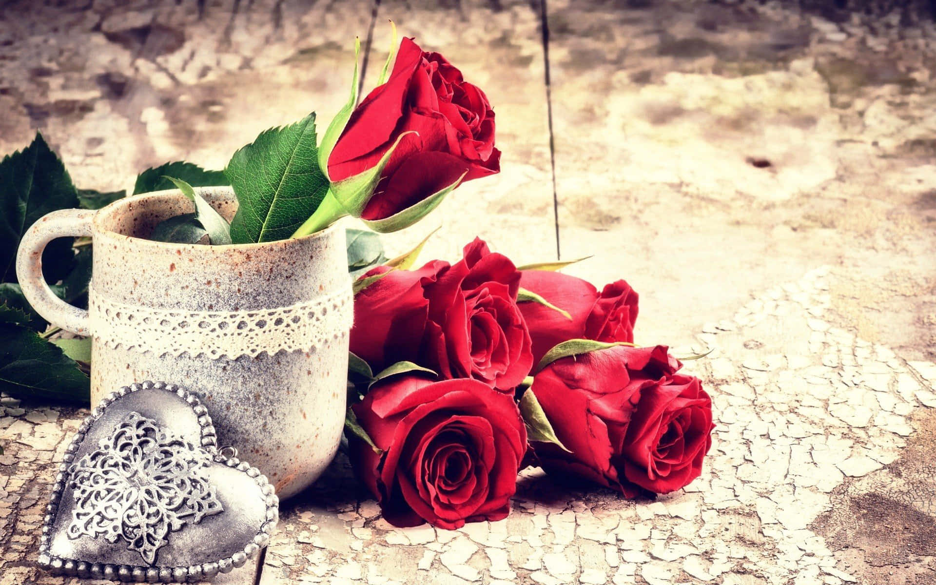 Rose Heart 桌布 壁紙 — 一個帶有蕾絲邊的鄉村風格陶瓷杯，在風化的木質表面上，旁邊放著一簇鮮豔的紅玫瑰和一個銀色鏤空心形，杯中插著一朵紅玫瑰。