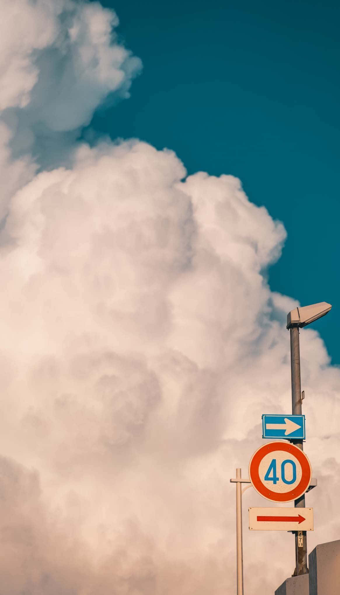 Minimalist Photography 桌布 壁紙 — 一個帶有速度限制和方向箭頭的街道標誌，矗立在戲劇性的天空下，天空中有蓬鬆的雲朵和藍綠色。