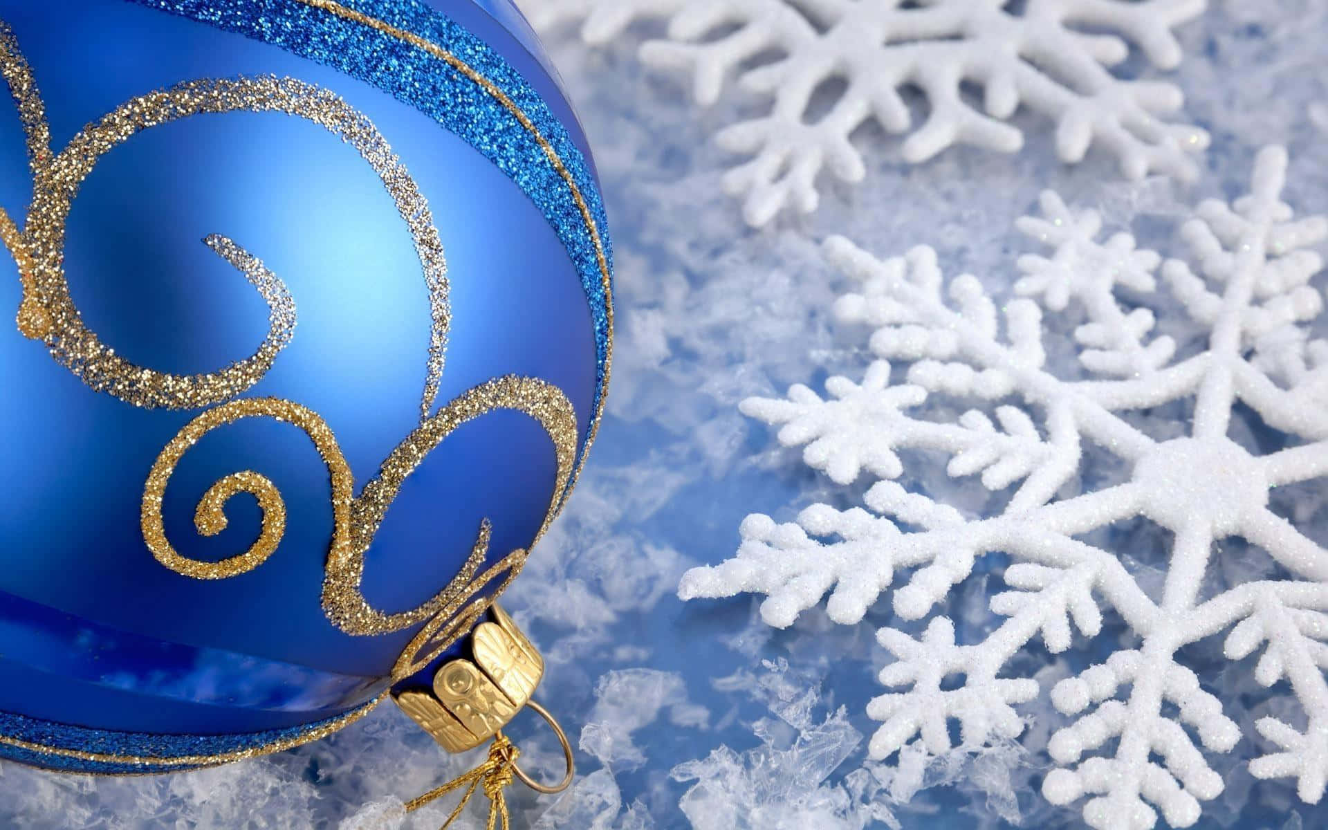 Winter Holiday 桌布 壁紙 — 一個帶有金色閃粉漩渦的光滑藍色聖誕裝飾品，放在白雪覆蓋的表面上，旁邊有白色雪花。