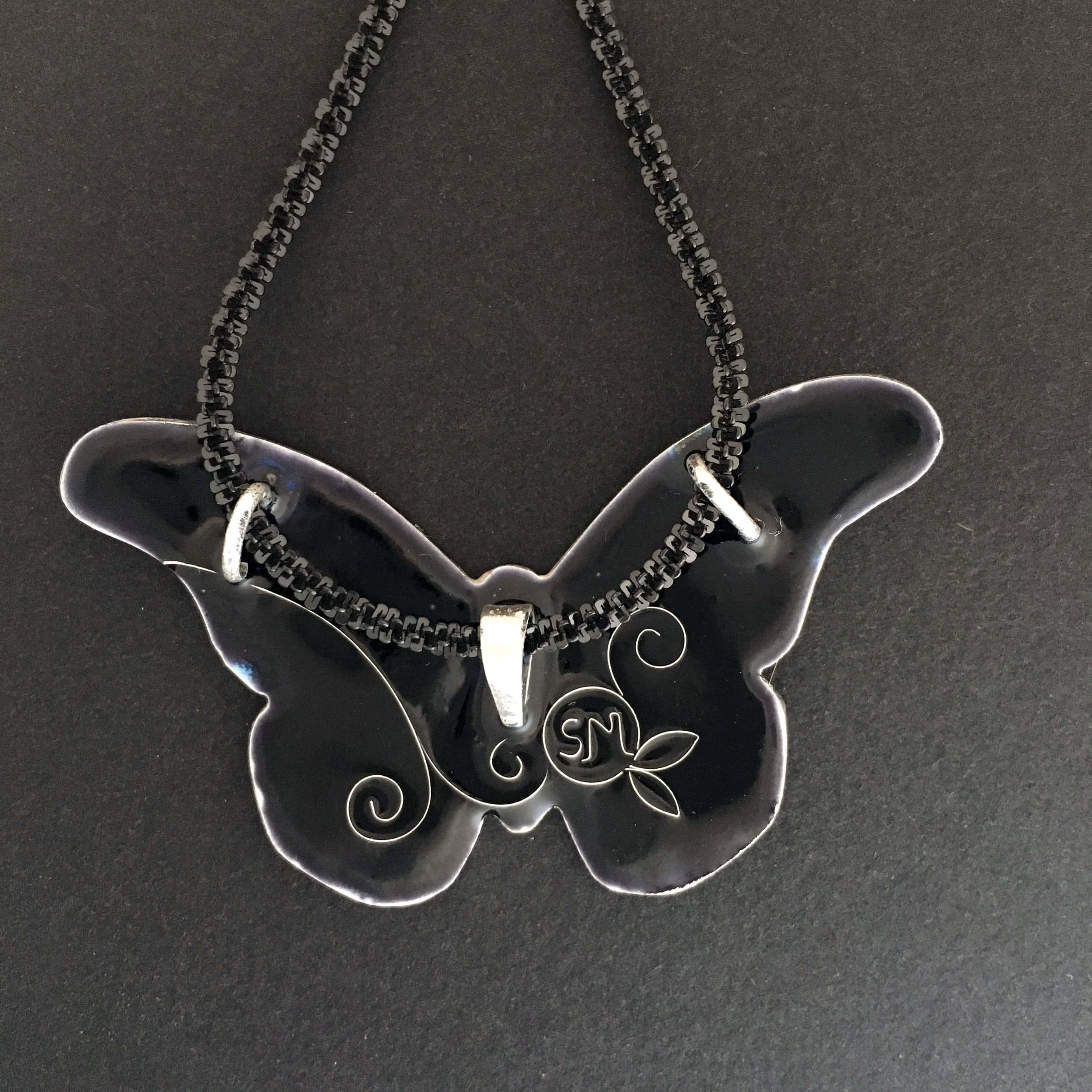 Butterfly Jewelry 桌布 壁紙 — 一個帶有銀色漩渦和字母組合的光澤黑色蝴蝶形吊墜，掛在珠鏈上，背景為深色紋理。