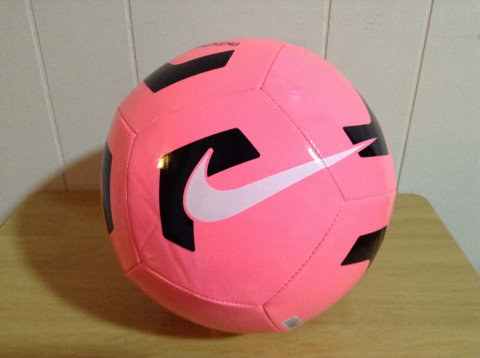 Pink Soccer Ball 桌布 壁紙 — 一個帶有黑色和白色點綴的鮮豔粉紅色Nike足球放在木質表面上，靠著淺色牆壁。