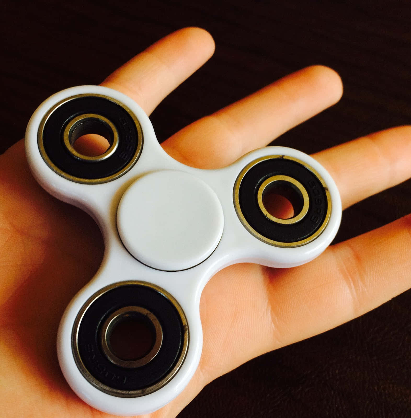 Fidget 桌布 壁紙 — 一個帶有黑色和金色軸承的白色指尖陀螺，放在張開的手掌上，背景係深色木質表面。