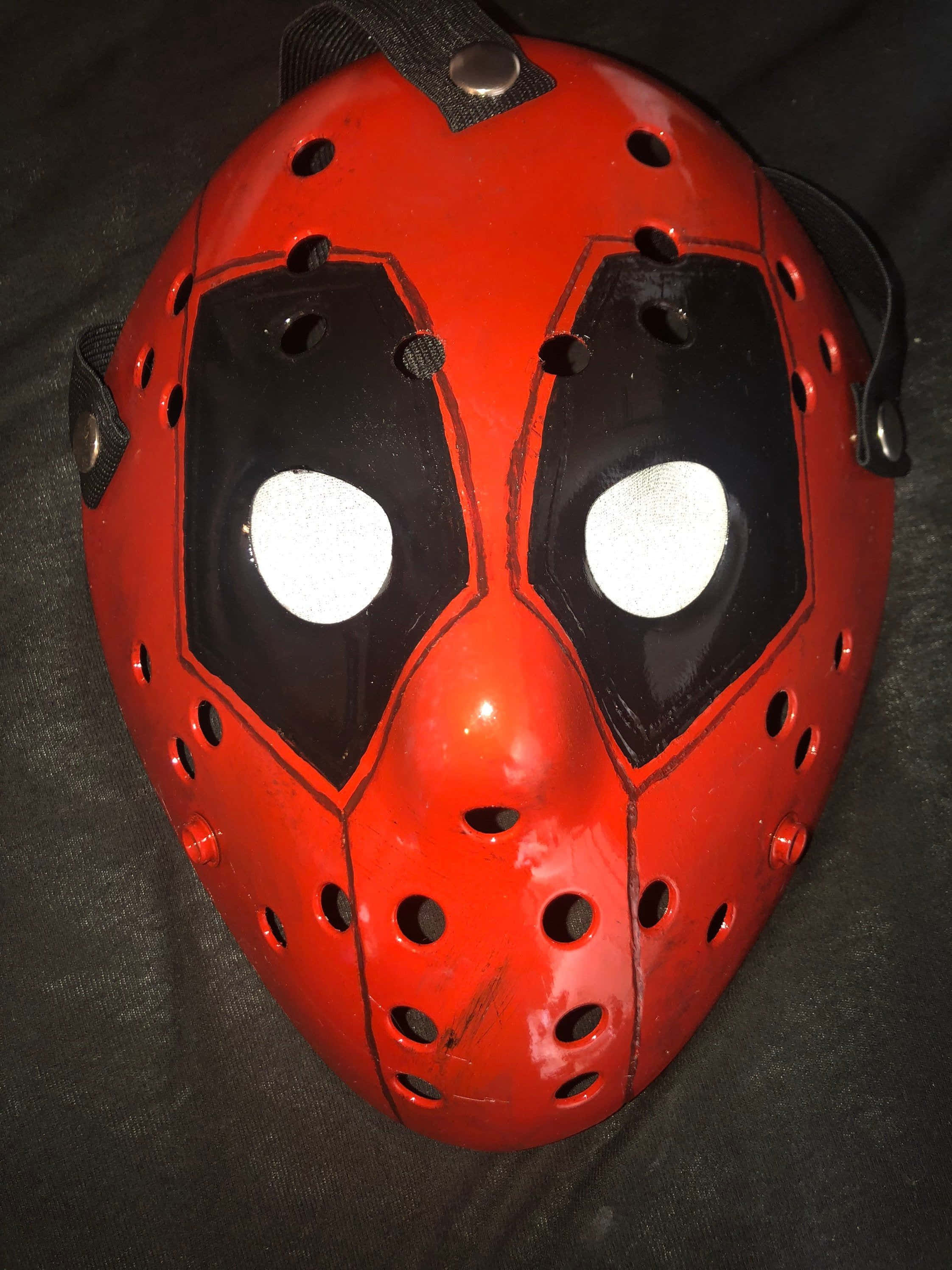 Deadpool Mask 桌布 壁紙 — 一個帶有黑色眼部鏤空和白色鏡片的亮面紅色曲棍球面具，放置在深色布料表面上。