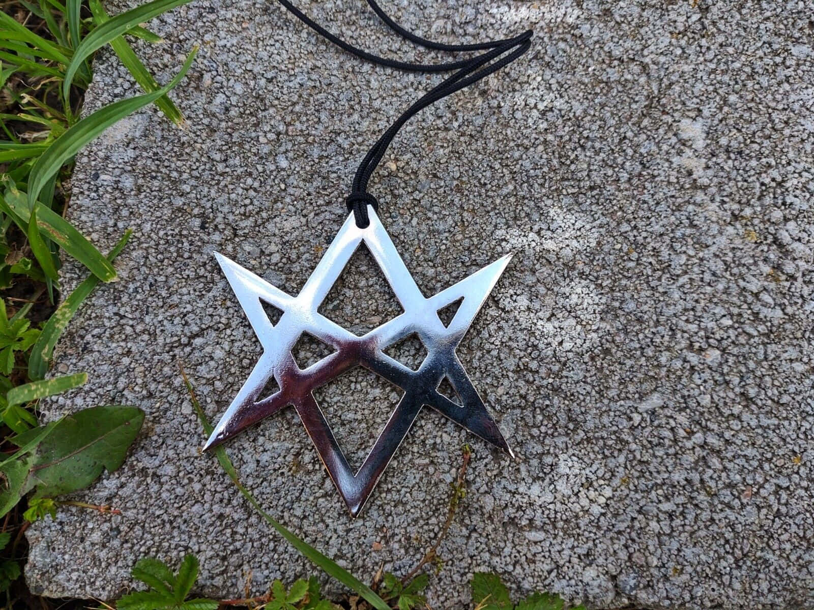 Thelema 桌布 壁紙 — 一個帶有黑色繩子的金屬五角星吊墜，放置在紋理混凝土上，旁邊是綠色草地。
