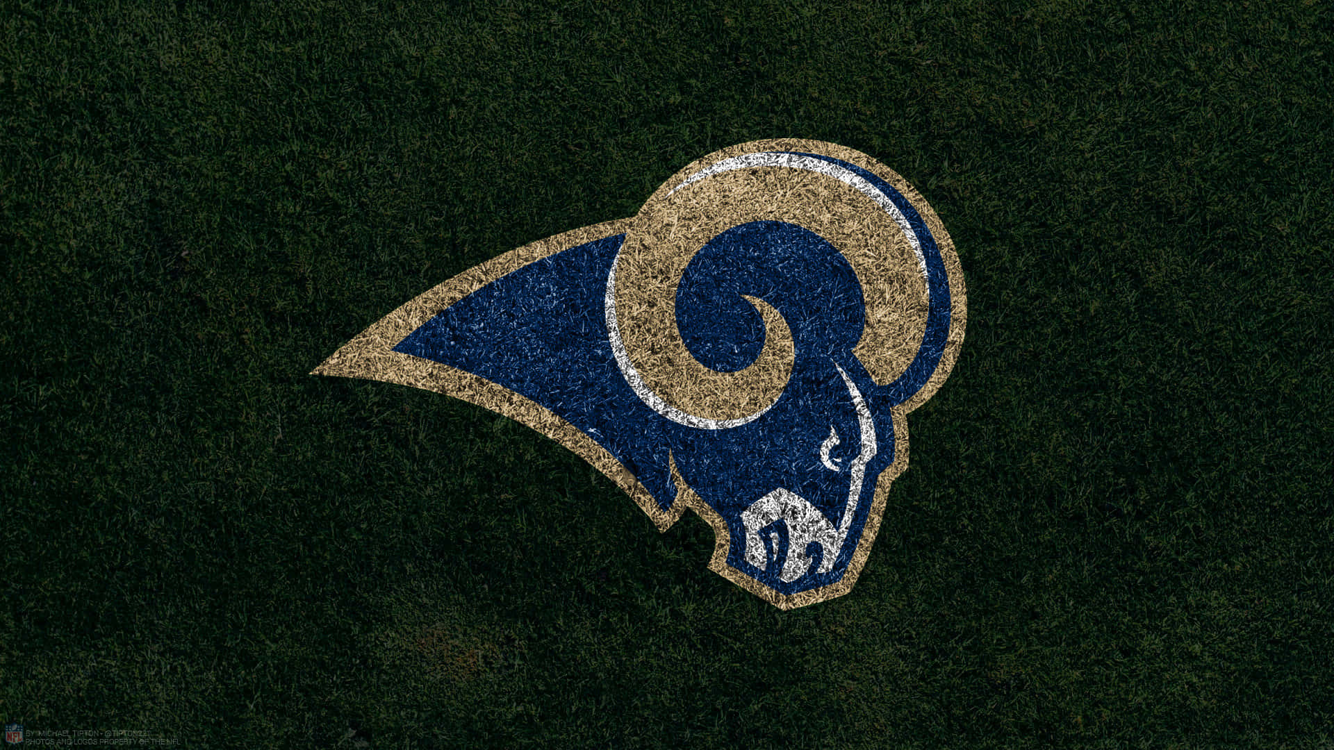 Rams 桌布 壁紙 — 一個帶紋理的公羊標誌，以藍色、金色和白色呈現，背景是深綠色的草地。
