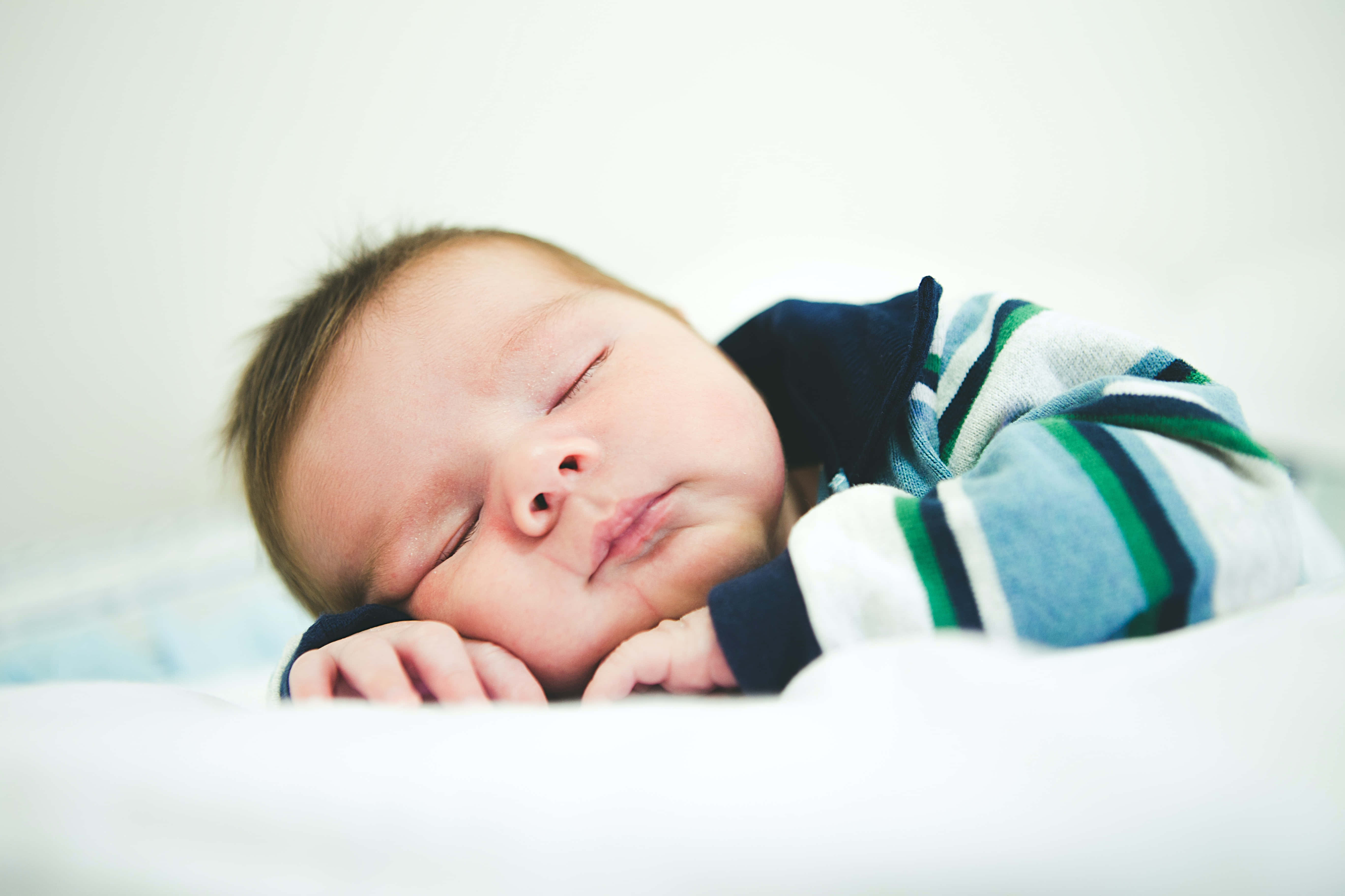 Baby Boy 桌布 壁紙 — 一個平靜的新生嬰兒側睡，包裹在柔軟的白色毛毯中，穿著藍、綠、白條紋連身衣，睡得香甜。