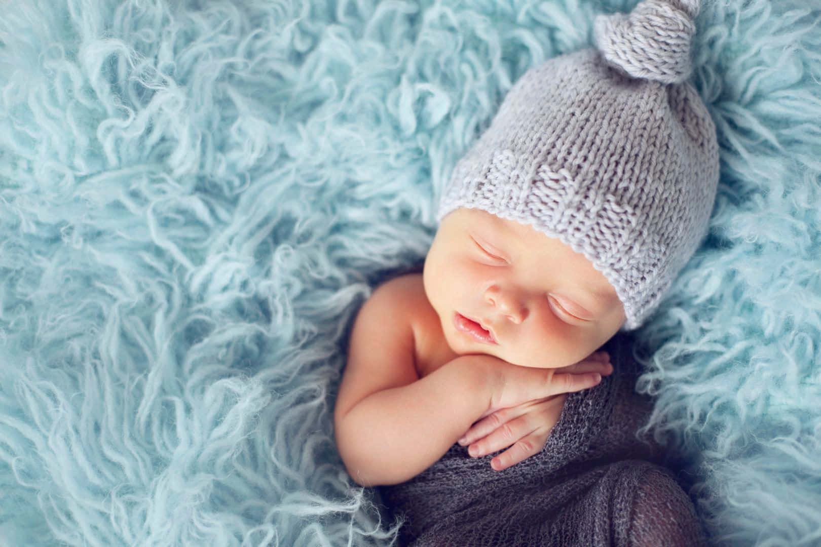Newborn Baby 桌布 壁紙 — 一個平靜的新生嬰兒戴著柔軟的灰色針織帽，在蓬鬆的淺藍色地毯上安睡。