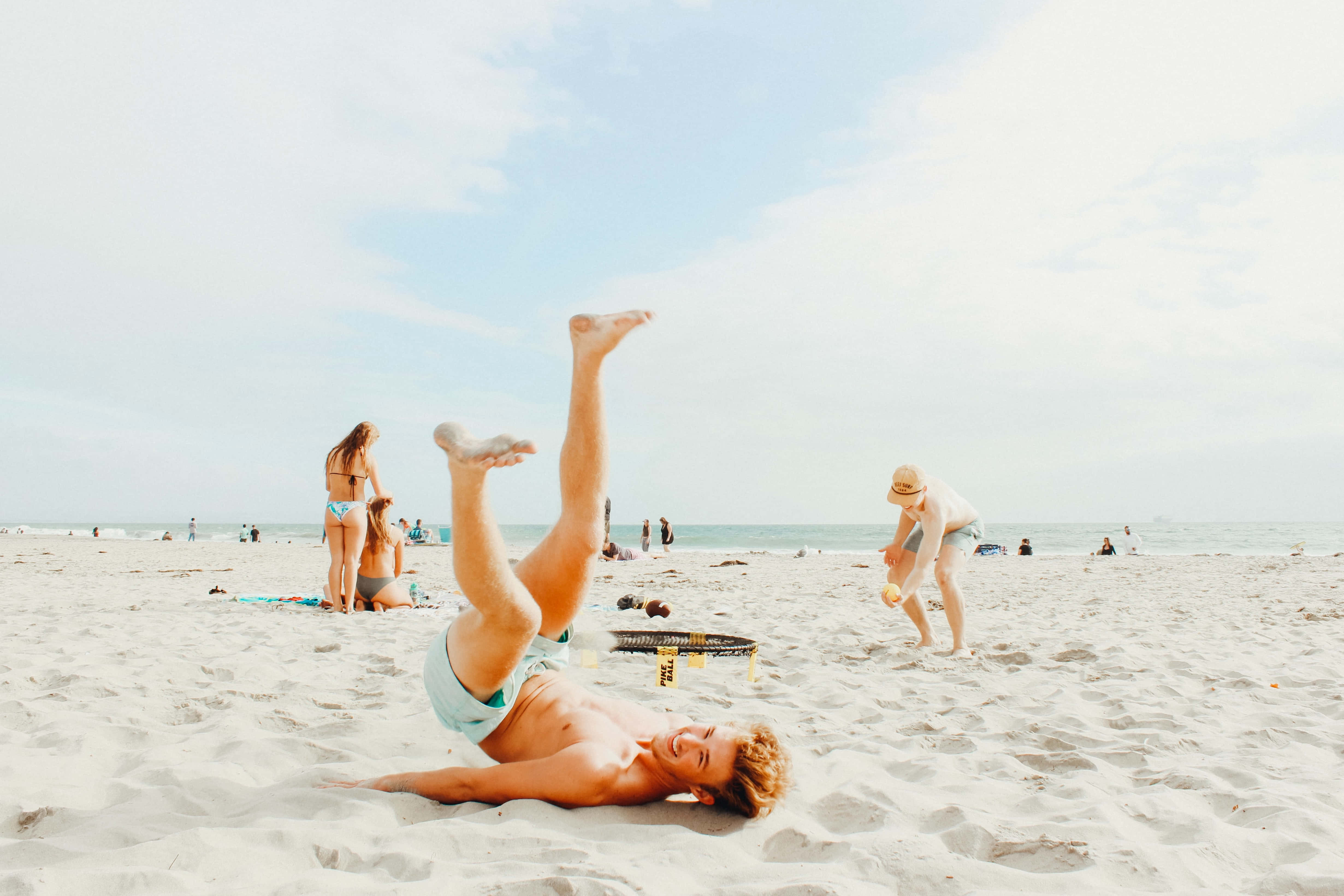 Summer Vacation 桌布 壁紙 — 一個年輕人在陽光明媚的海灘上嬉戲地倒立，笑著，而其他人在附近放鬆。