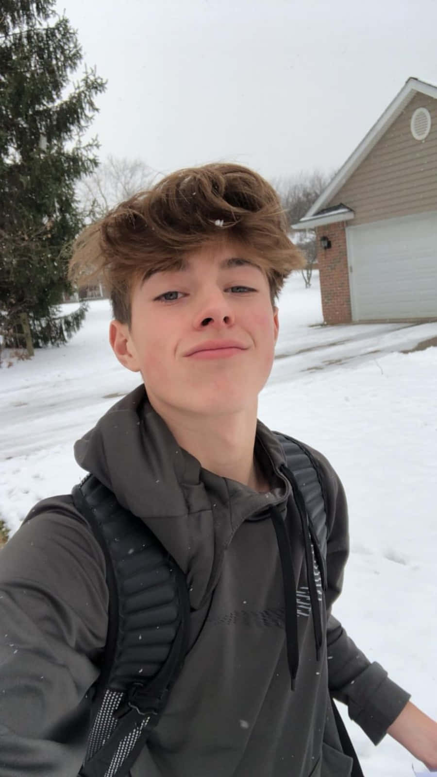 Boy 桌布 壁紙 — 一個年輕人在雪覆蓋的郊區庭院中，頭髮蓬鬆，背著背包，溫柔地微笑。