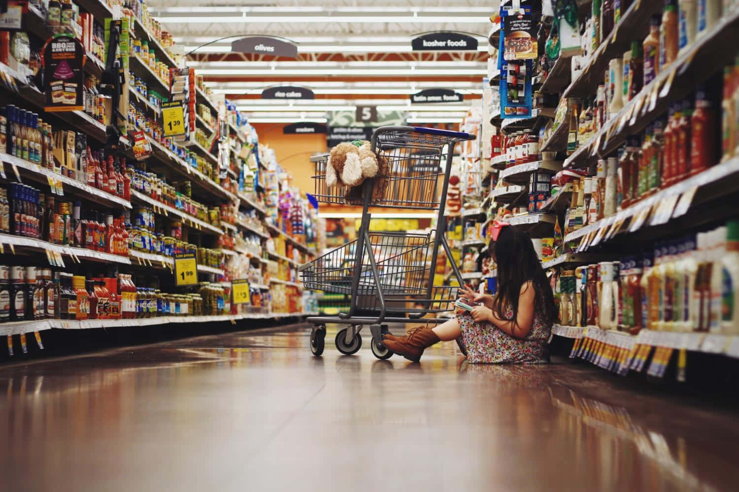 Grocery Store 桌布 壁紙 — 一個年輕女孩坐在超市地板上，旁邊是裝滿雜貨的購物車，周圍是明亮的螢光燈下高聳的產品貨架。