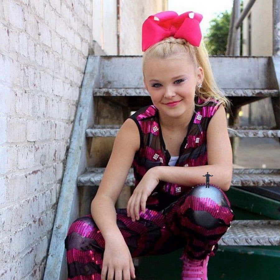jojo siwa 桌布 壁紙 — 一個年輕女孩戴著一個巨大的粉紅色蝴蝶結，坐在金屬樓梯上，背景是白色磚牆，身穿閃亮的粉紅色和黑色服裝。