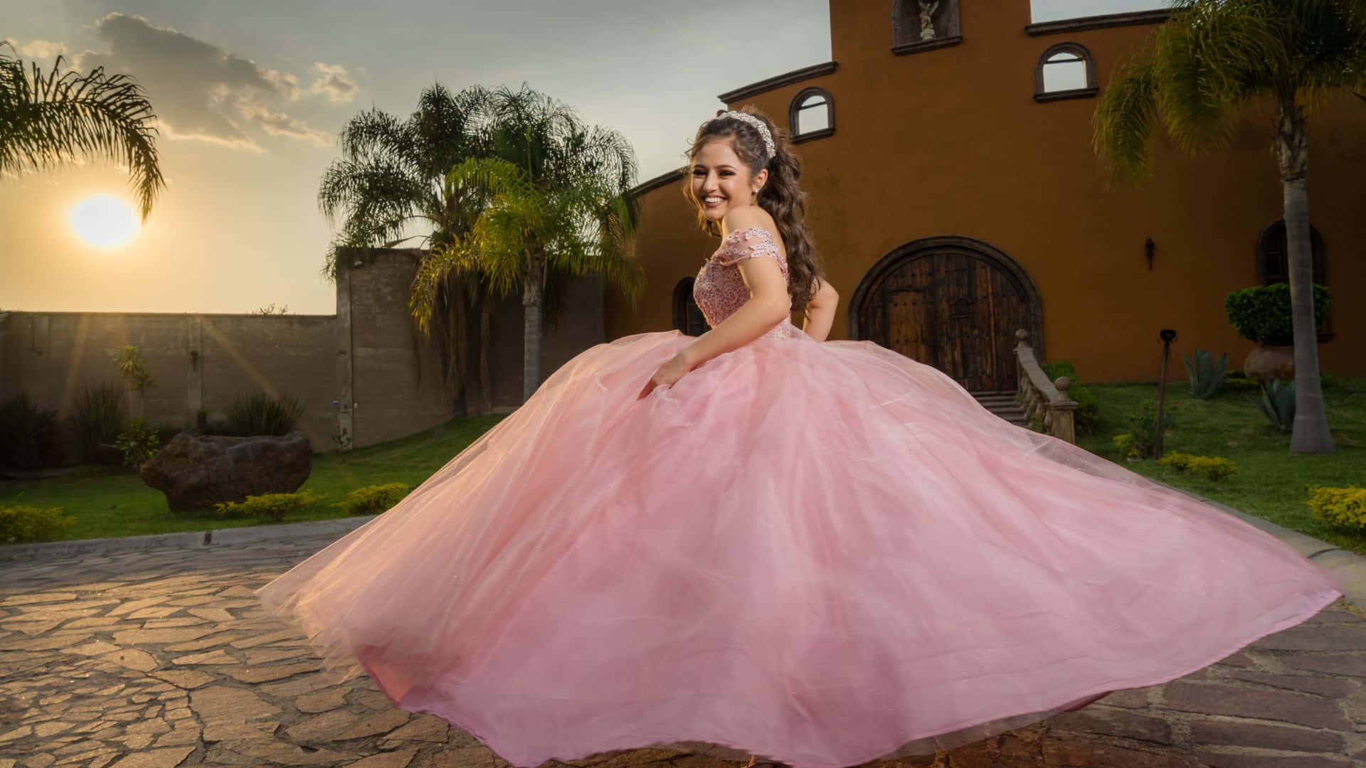 Pink Dress 桌布 壁紙 — 一個年輕女孩穿著飄逸的粉紅色 quinceanera 禮服，在石頭庭院裡快樂地旋轉，夕陽落在棕櫚樹後。