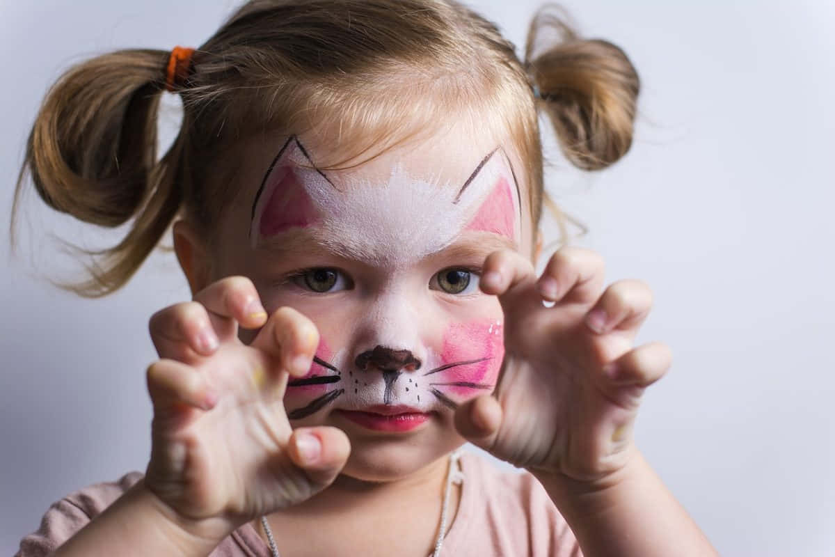 Halloween Face Paint 桌布 壁紙 — 一個年輕女孩臉上畫著貓咪彩繪，紮著雙馬尾，在素色背景前頑皮地將手靠近臉龐擺姿勢。