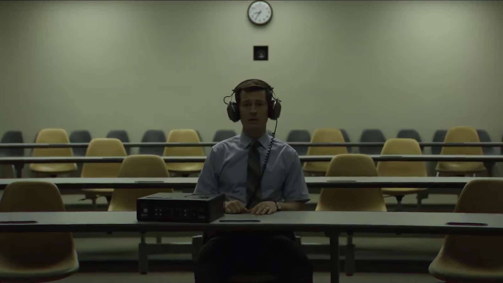 Caption: Unforgettable Scenes from the Mindhunter Series 壁紙 — 一個年輕男子獨自坐在教室裏，戴着耳機，桌上放着一部復古音頻設備，上方有掛牆時鐘。