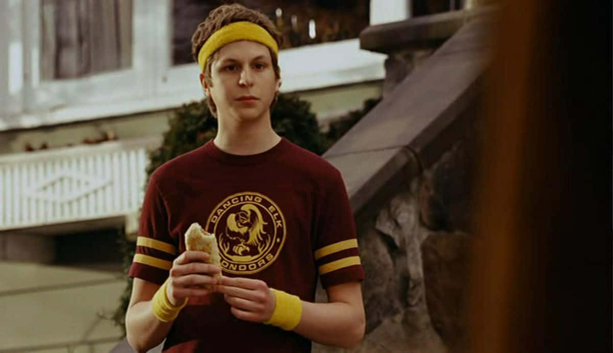michael cera 桌布 壁紙 — 一個年輕男子穿著栗色 'Dancing Elk' T恤和黃色頭帶，站在屋外的石階上，手裡拿著一個三文治。