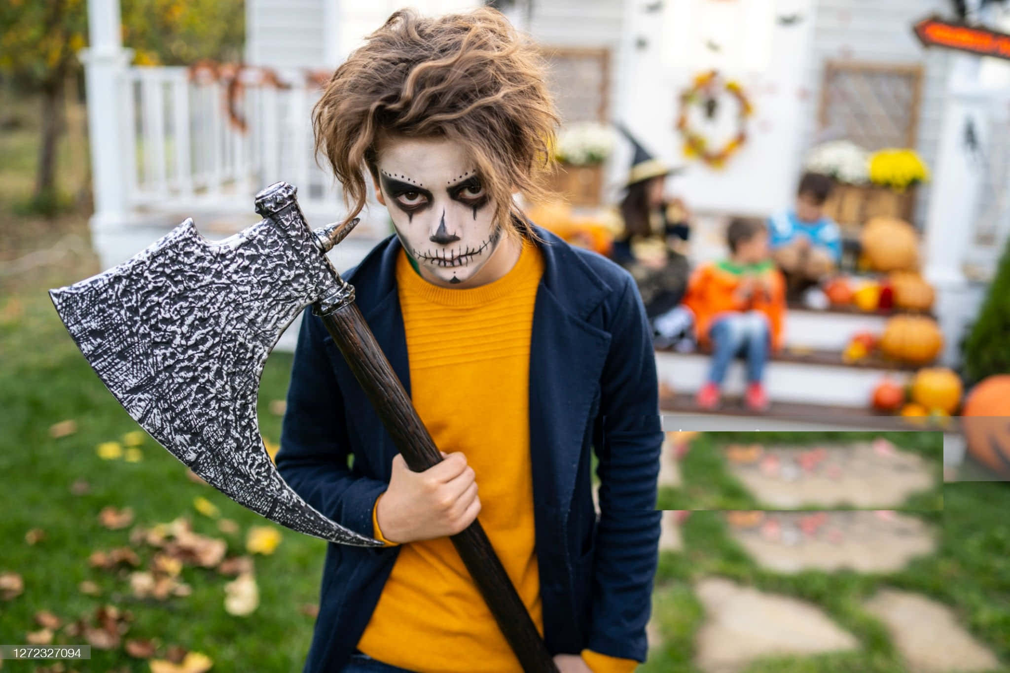 Zombie Costumes 桌布 壁紙 — 一個年輕男孩戴著骷髏化妝，拿著一把大斧頭，在裝飾著南瓜和秋葉的門廊上玩不給糖就搗蛋。