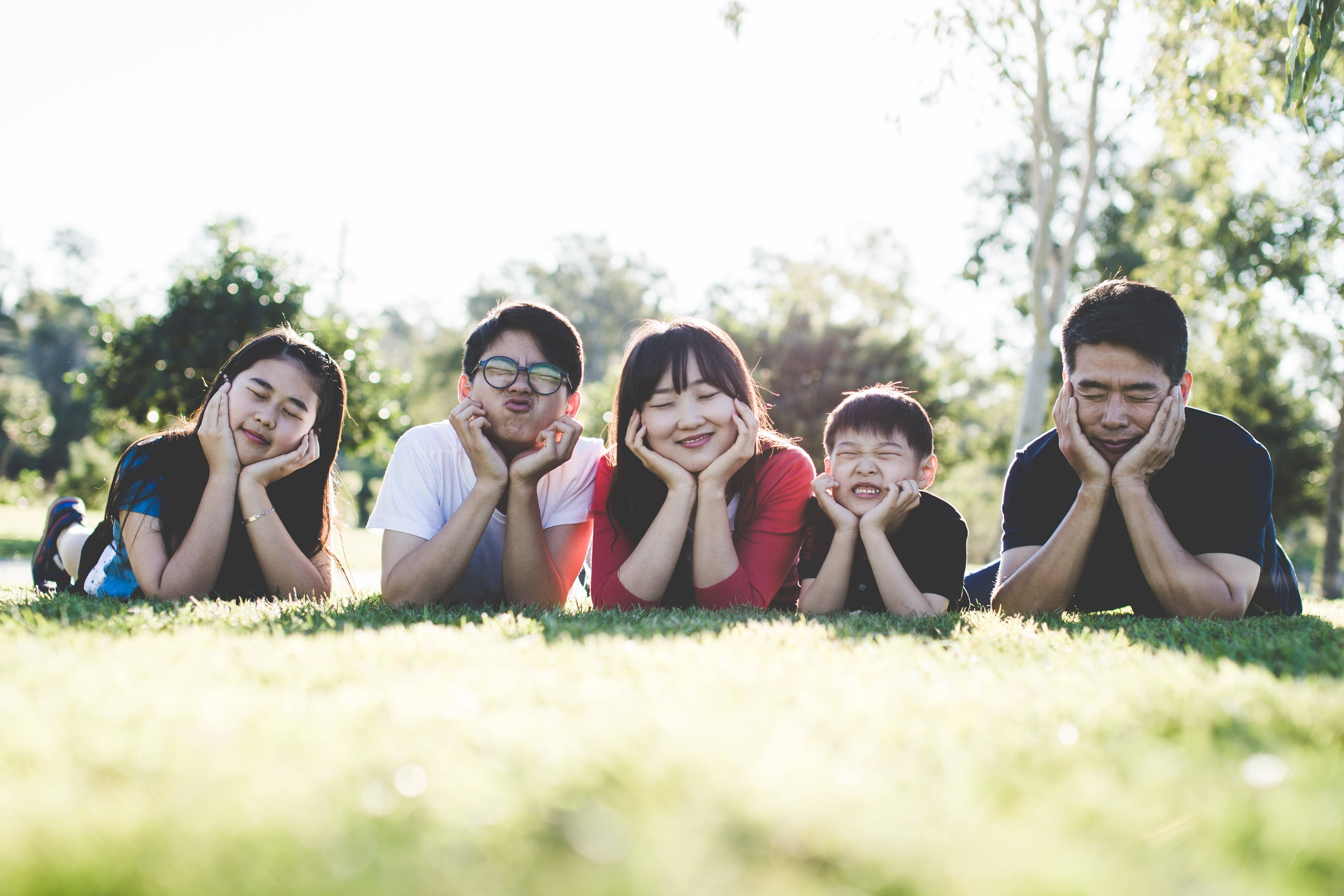 Family 桌布 壁紙 — 一個幸福的五口之家躺在陽光普照的草地上，在公園裡頑皮地將手放在下巴下擺姿勢。