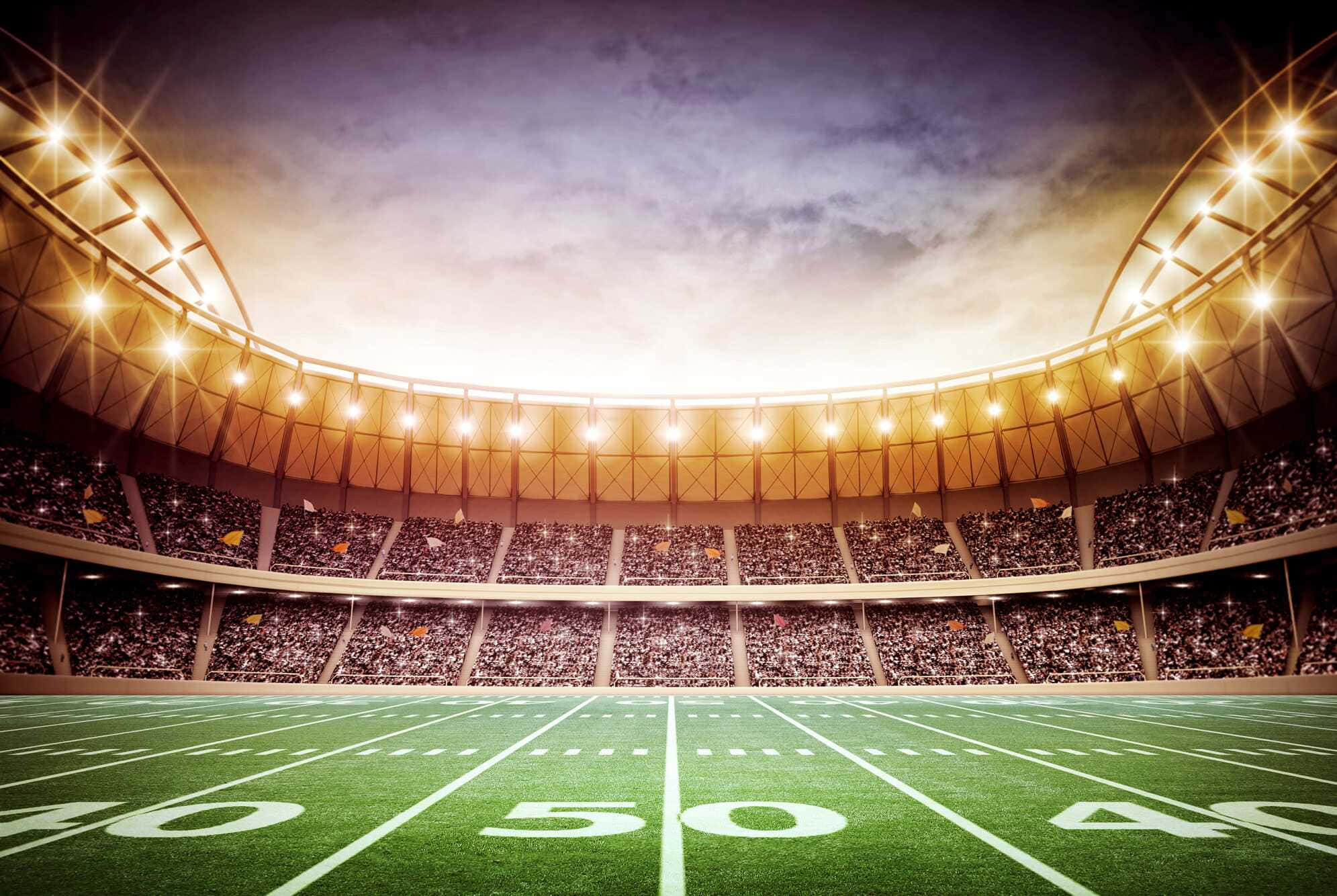 Super Bowl 桌布 壁紙 — 一個廣闊的足球場，在黃金時段閃爍的泛光燈下，擠滿了歡呼的球迷。