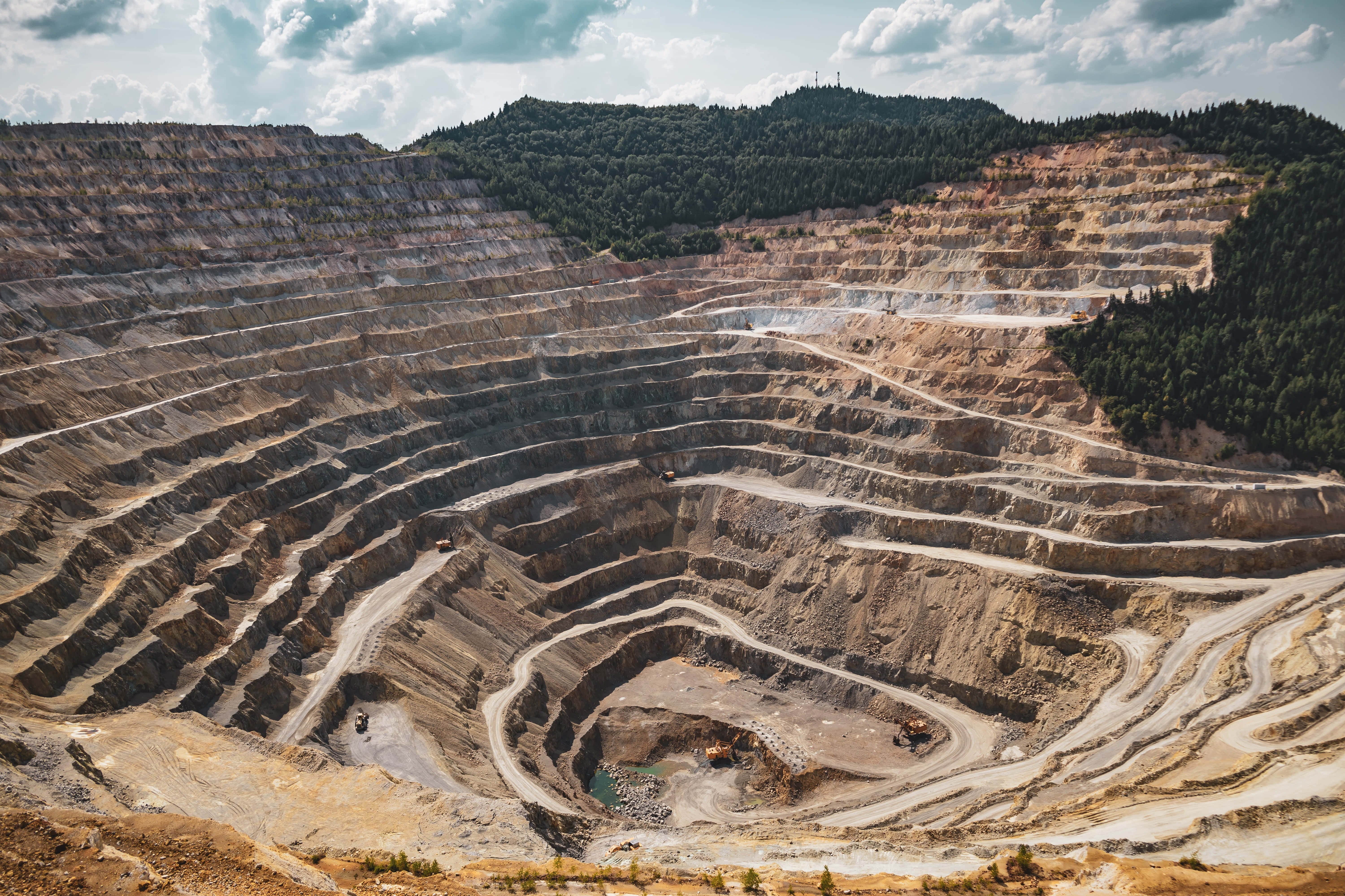 Mining 桌布 壁紙 — 一個廣闊的露天礦場，擁有梯田狀的土牆，天空部分多雲，周圍是森林覆蓋的山丘。