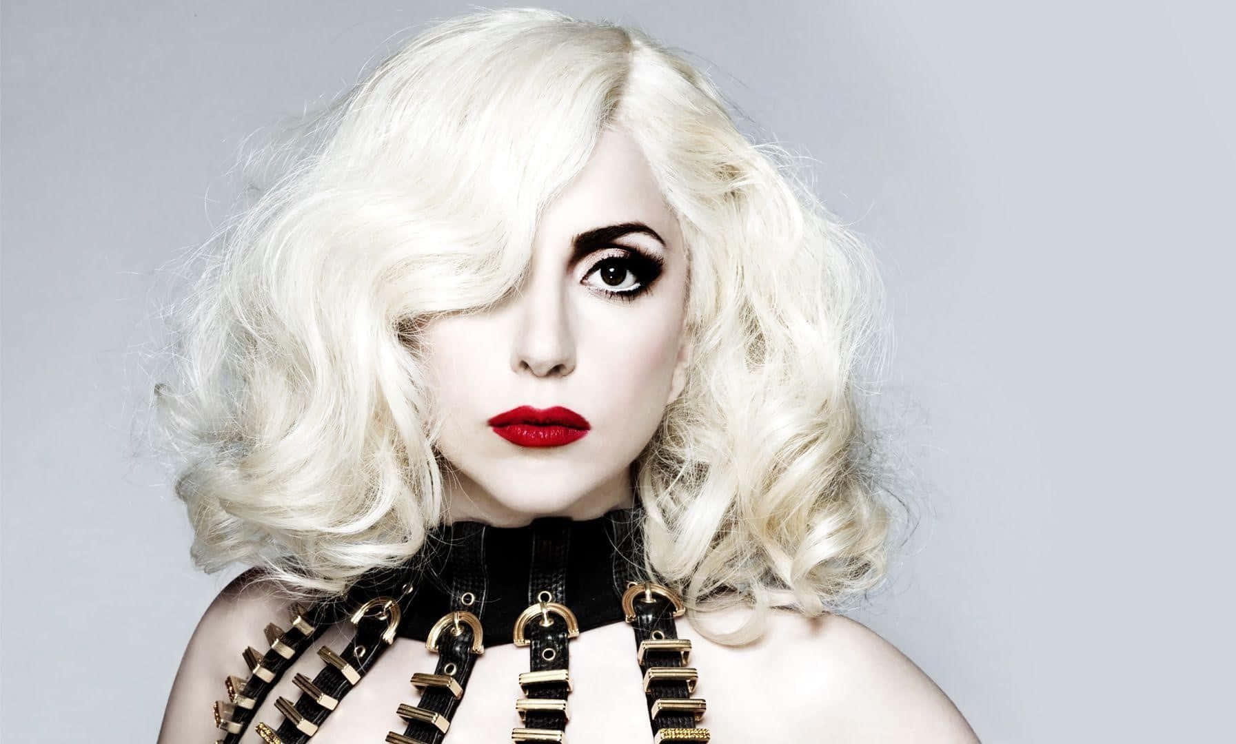 Lady Gaga 桌布 壁紙 — 一個引人注目的女性肖像，擁有鉑金色捲髮、大膽的紅唇和黑色皮革背帶，背景是純灰色。
