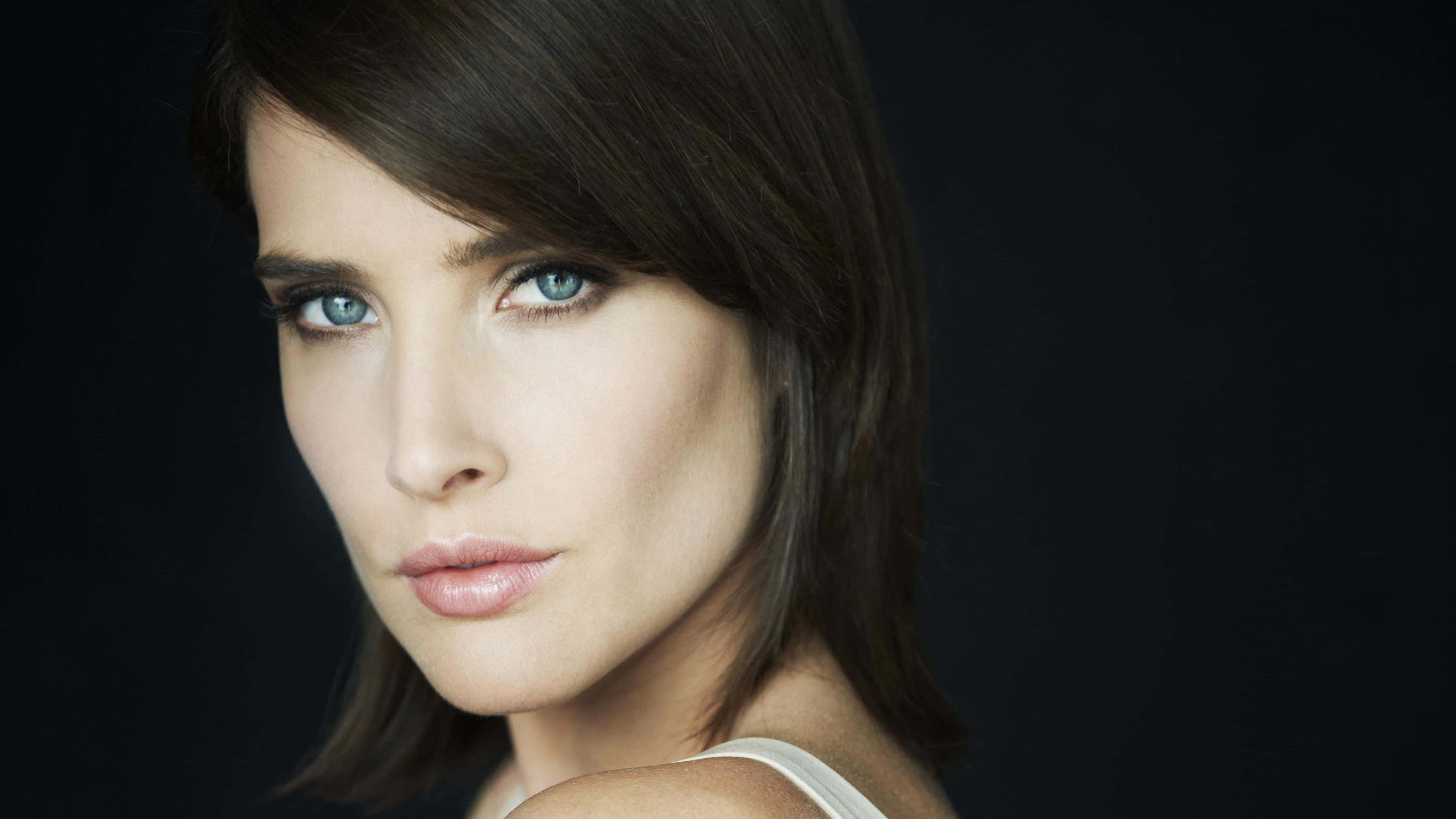 Cobie Smulders 桌布 壁紙 — 一個引人注目的肖像，一位擁有銳利藍眼睛和深色頭髮的女性，背景為黑色。
