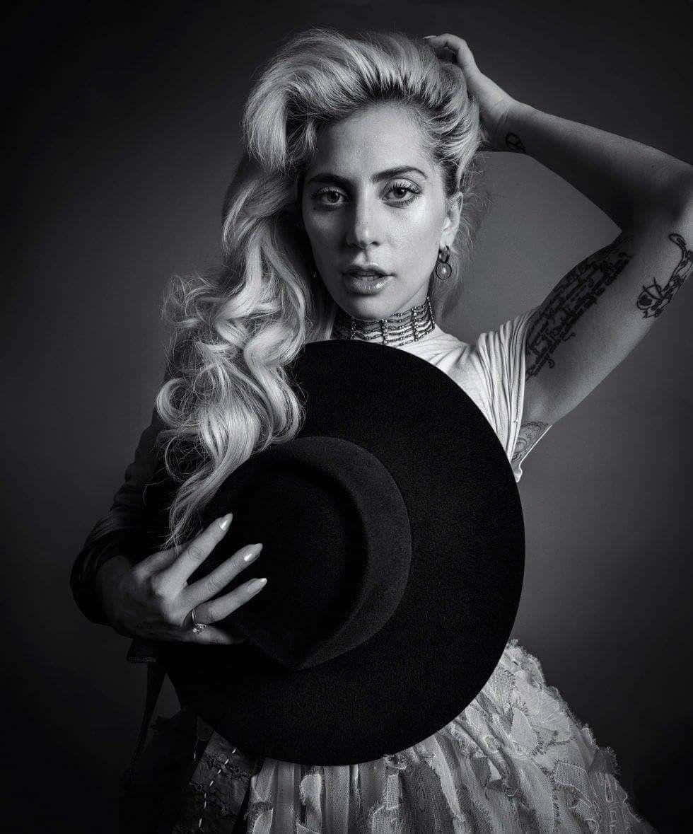 Lady Gaga 桌布 壁紙 — 一個引人注目的黑白肖像，一位擁有豐盈金色捲髮的女性在深色攝影棚背景下拿著一頂寬邊帽。