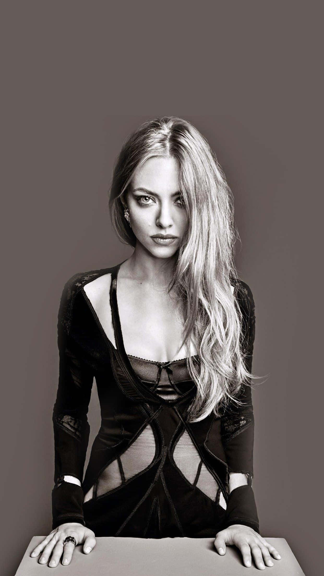 Amanda Seyfried 桌布 壁紙 — 一個引人注目的黑白肖像，一位長金髮女性穿著暴露的黑色連衣裙靠在桌子上。