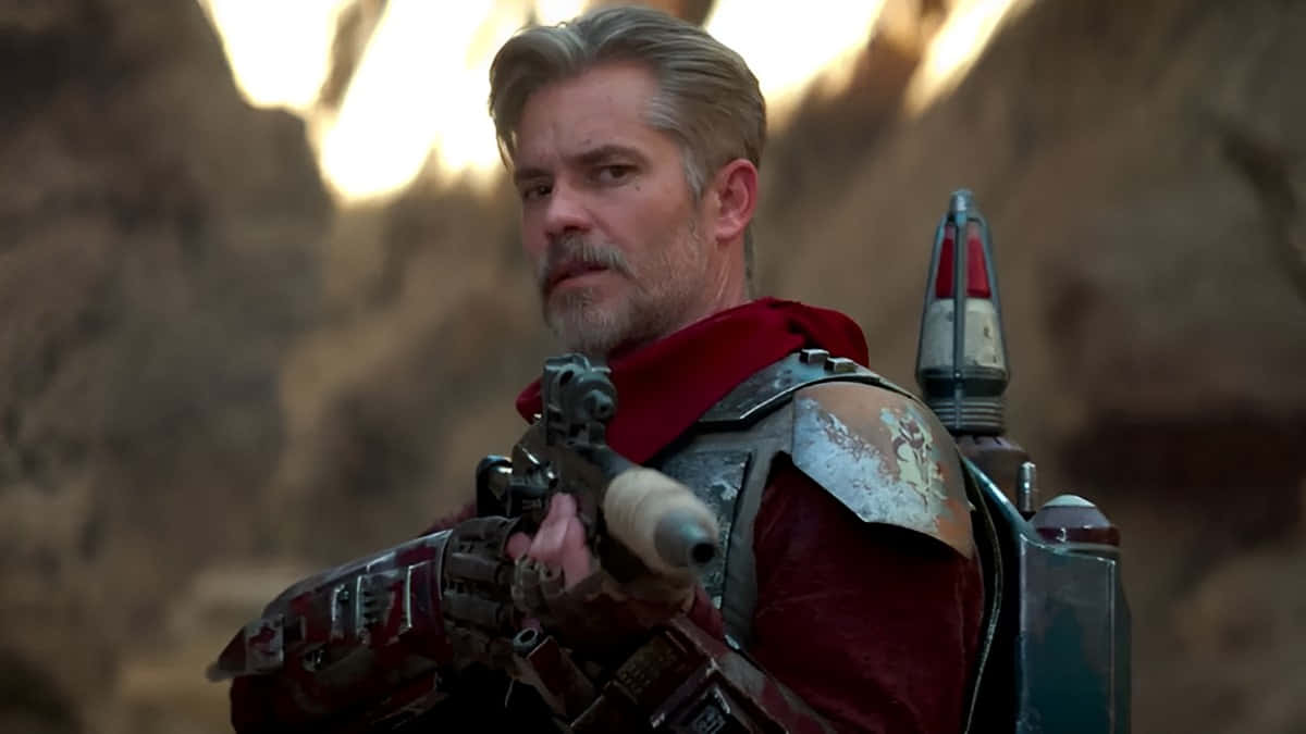timothy olyphant 桌布 壁紙 — 一個強悍的賞金獵人身穿紅色盔甲，在岩石峽谷中保持警覺，身後是溫暖的夕陽。