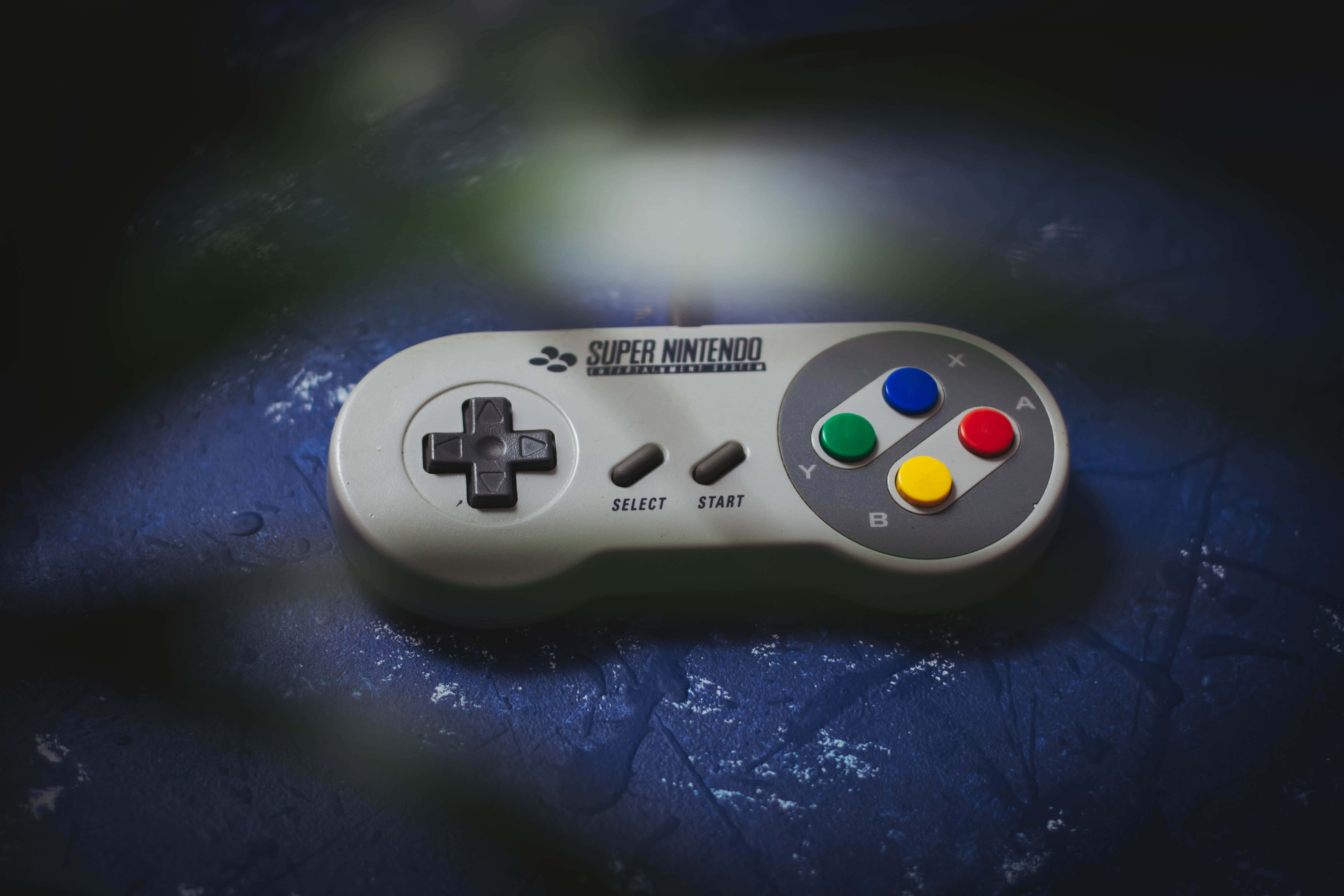 Home Entertainment 桌布 壁紙 — 一個復古的Super Nintendo控制器放在有紋理的藍色表面上，在戲劇性的聚光燈下。