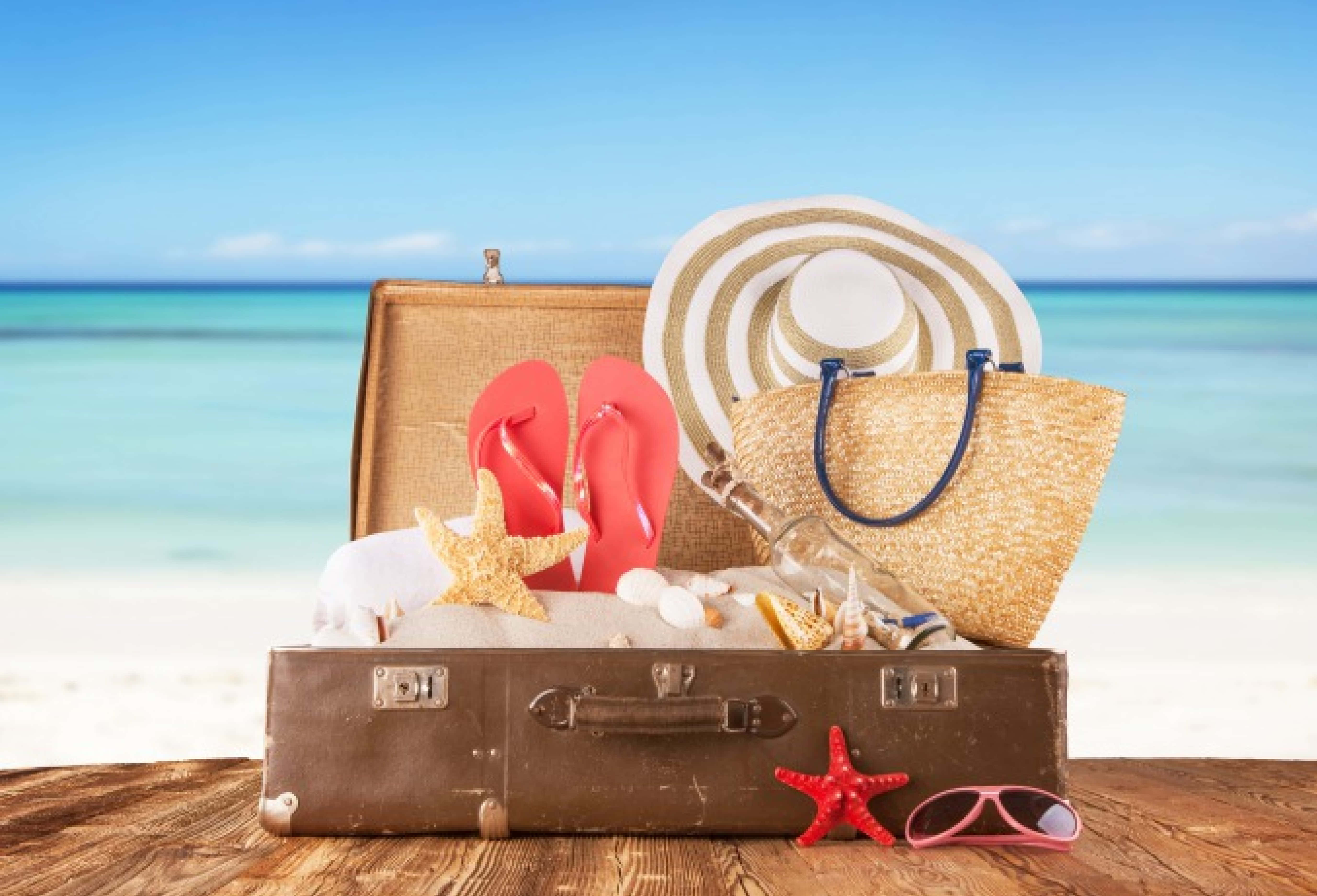 Beach Bag 桌布 壁紙 — 一個復古行李箱裝滿沙灘用品，放在木桌上，背景是藍綠色海洋。