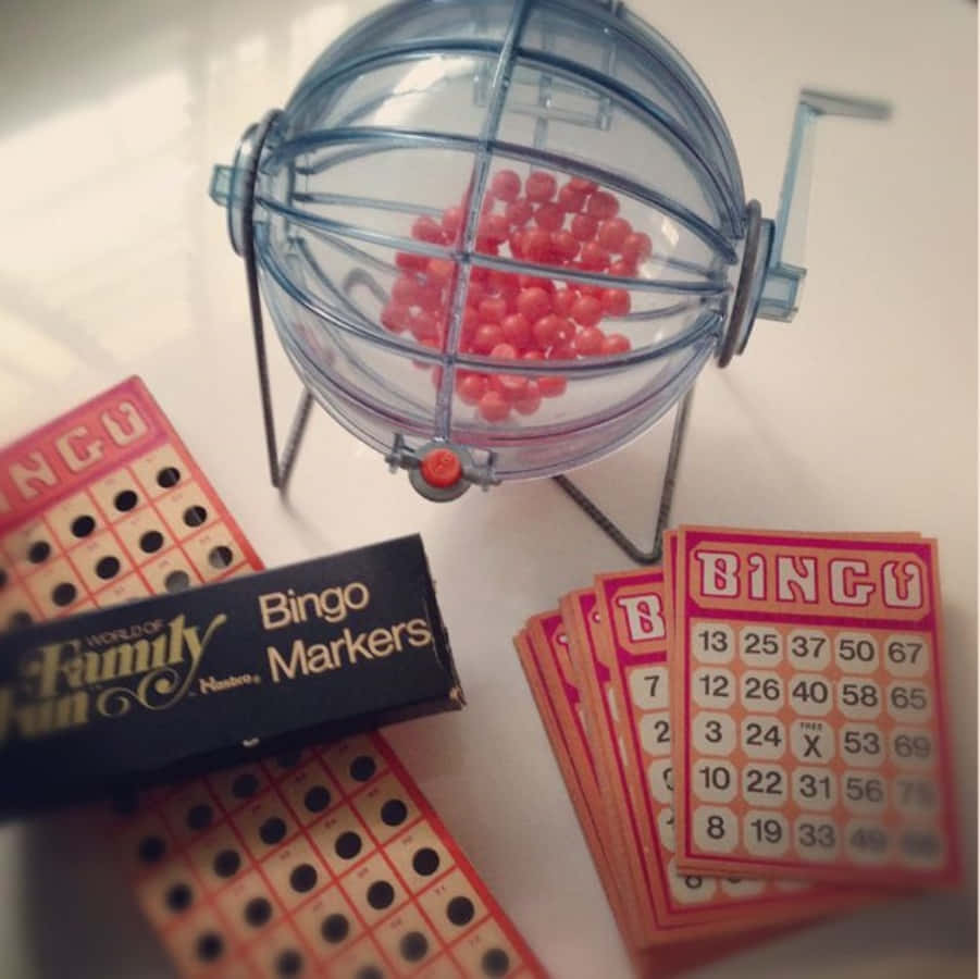 Bingo 桌布 壁紙 — 一個復古賓果設置，有一個透明球裝滿紅色籌碼，周圍是橙色卡片和一個黑色記號筆盒，放在白色表面上。