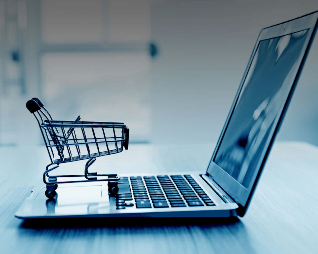 E Commerce 桌布 壁紙 — 一個微型購物車放在手提電腦鍵盤上，處於冷色調藍色單色辦公室環境中。