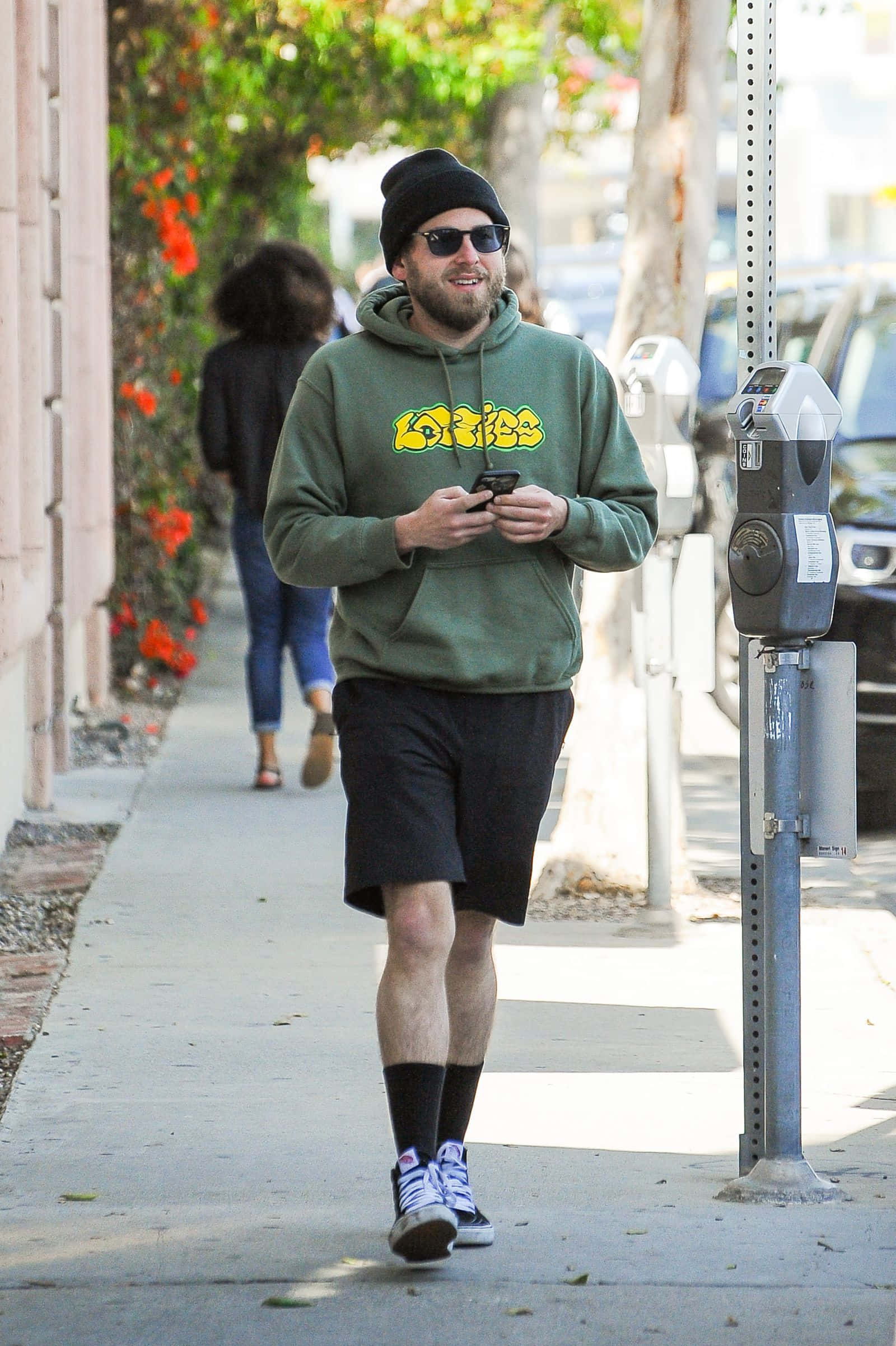 jonah hill 桌布 壁紙 — 一個微笑嘅男人，著住綠色連帽衫同冷帽，喺陽光普照嘅行人路上邊行邊睇手機。