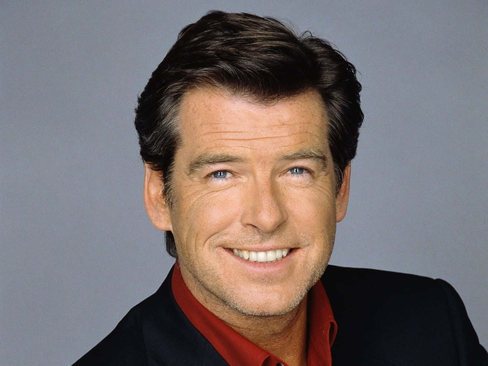 pierce brosnan 桌布 壁紙 — 一個微笑嘅男人嘅特寫肖像，佢有藍色眼睛，著住紅色恤衫同黑色外套，背景係灰色。
