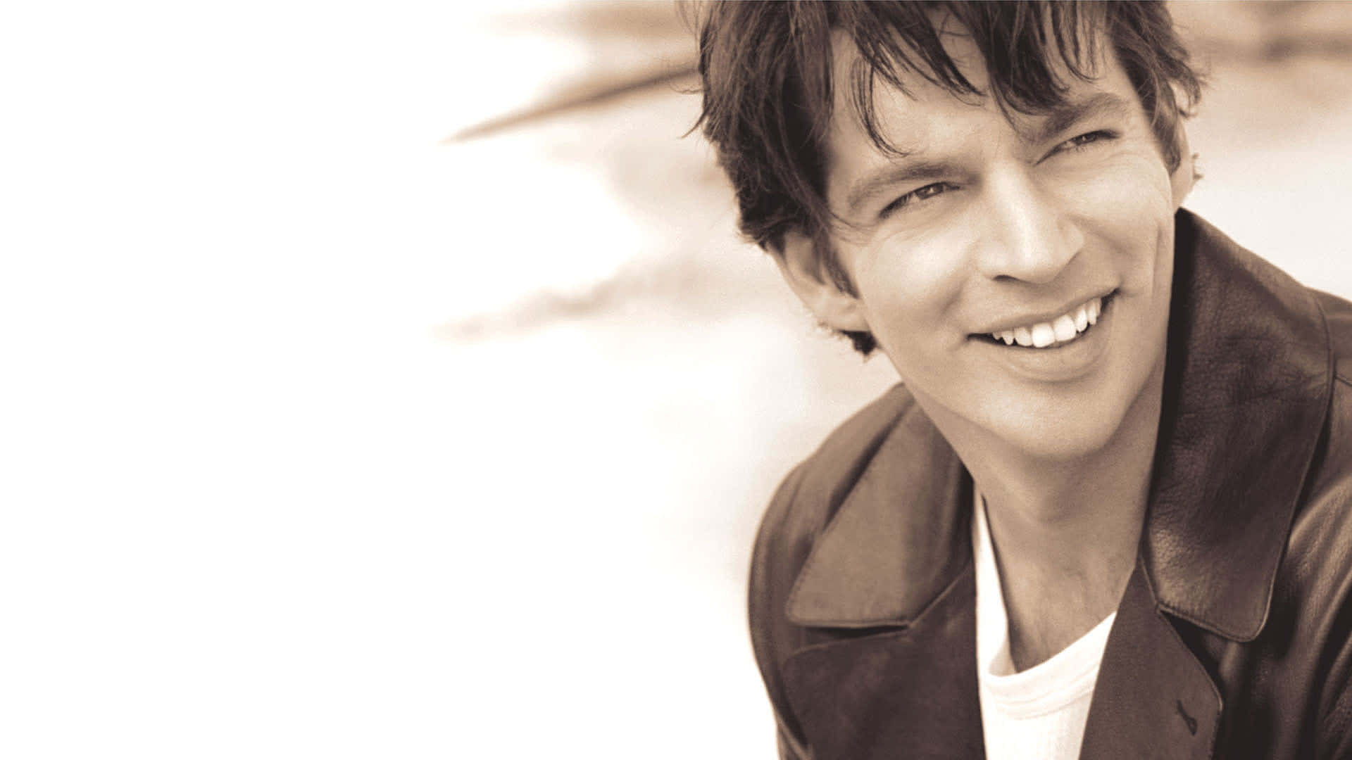 Harry Connick Jr 桌布 壁紙 — 一個微笑嘅男人著住皮褸，喺明亮模糊嘅背景下溫暖咁望住鏡頭。