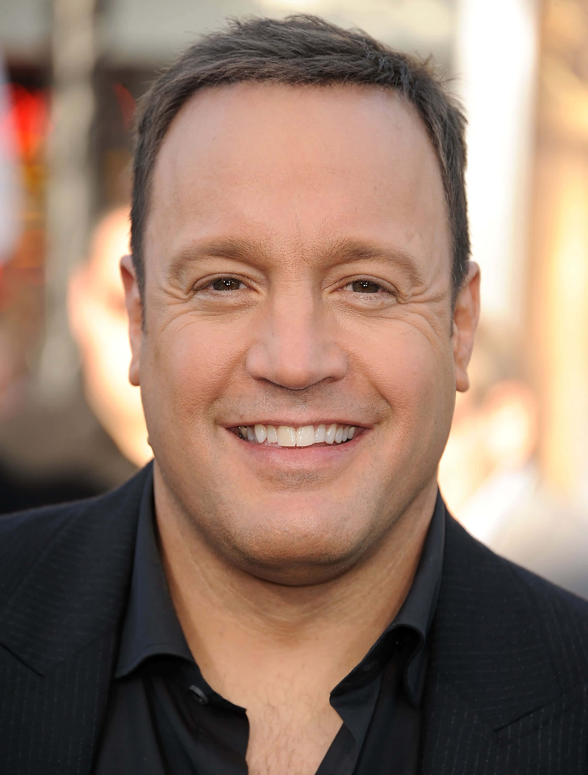 Kevin James 桌布 壁紙 — 一個微笑嘅短黑髮男人嘅特寫肖像，穿著黑色西裝同恤衫，背景係模糊嘅城市景觀。