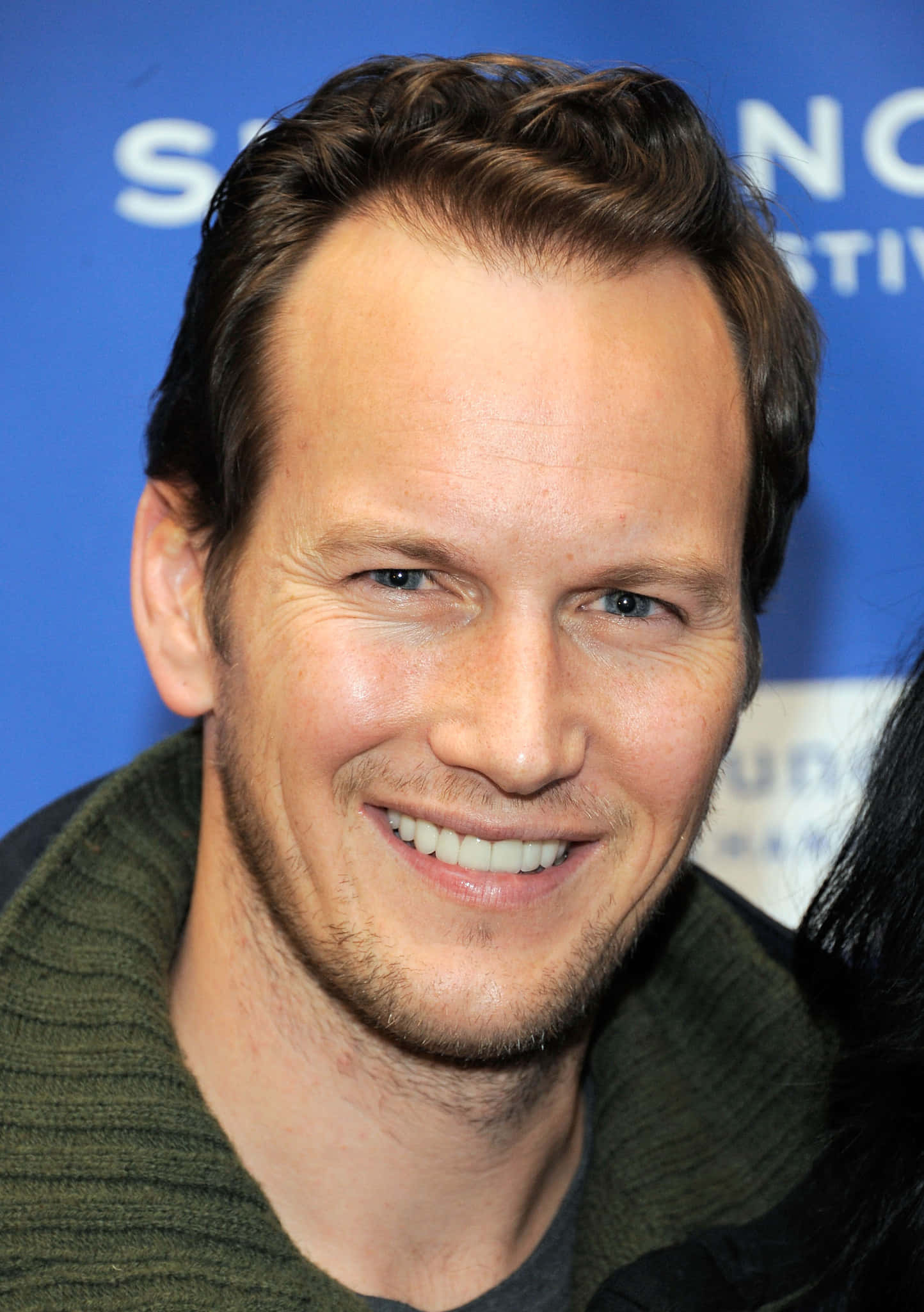 Patrick Wilson 桌布 壁紙 — 一個微笑男士的特寫照片，短棕髮和鬍渣，穿著綠色毛衣，背景是藍色活動背景。