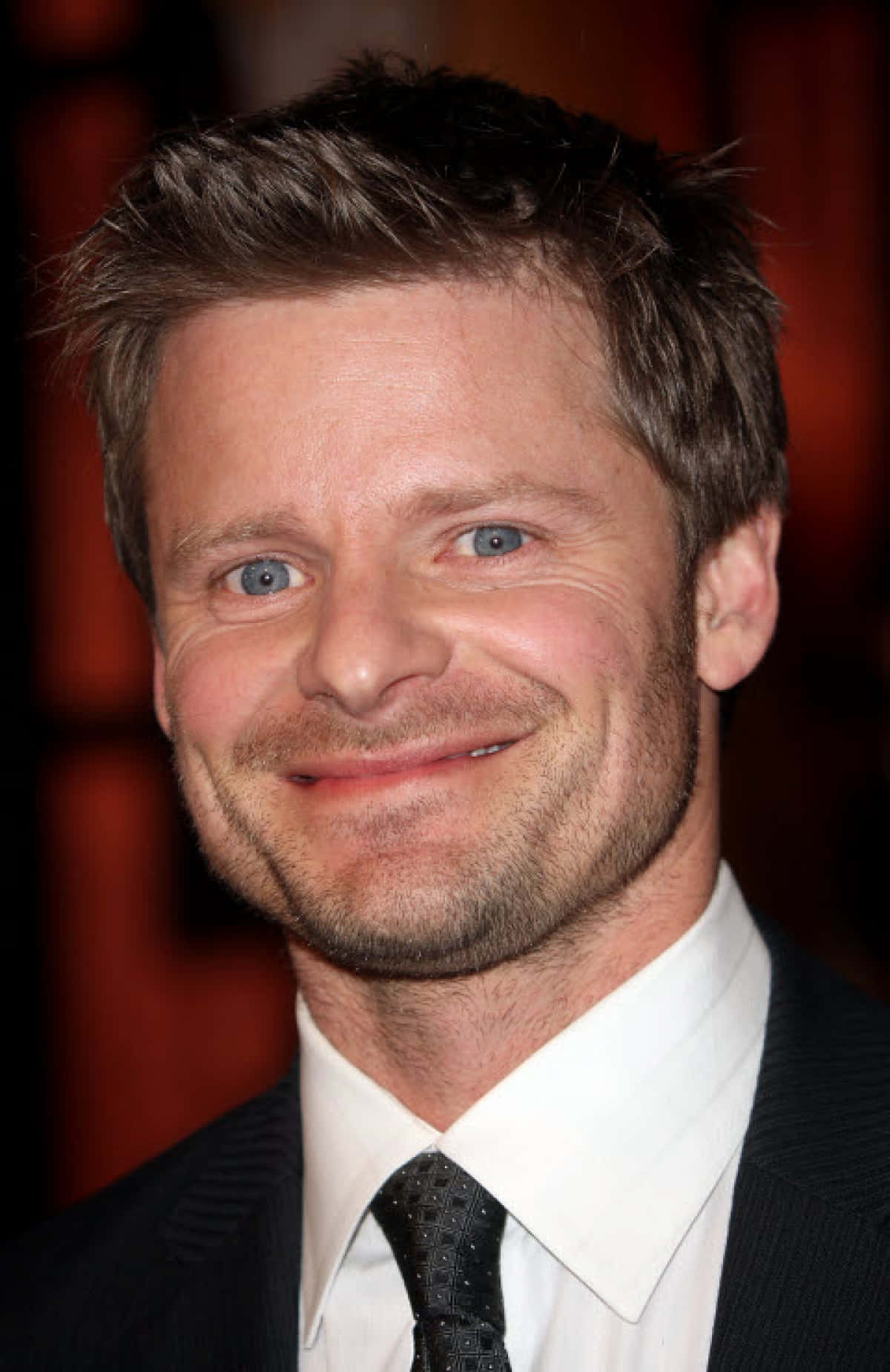 Steve Zahn 桌布 壁紙 — 一個微笑男士的特寫肖像，藍眼睛，鬍渣，穿著深色西裝和領帶，背景是模糊的紅色。