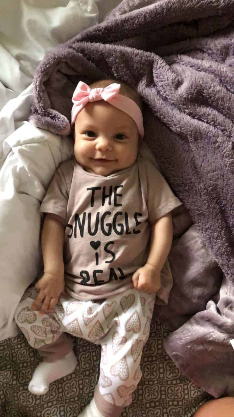 Baby Girl 桌布 壁紙 — 一個微笑的女嬰，戴著粉紅色蝴蝶結，穿著心形圖案褲子，躺在柔軟的紫色毯子上，穿著一件寫著「THE SNUGGLE IS REAL」的襯衫。