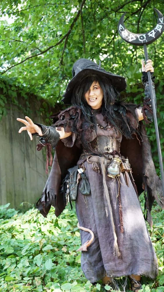 Witch Costumes 桌布 壁紙 — 一個微笑的女巫穿著破舊的深色服裝，站在陽光明媚的花園中，手持新月法杖。