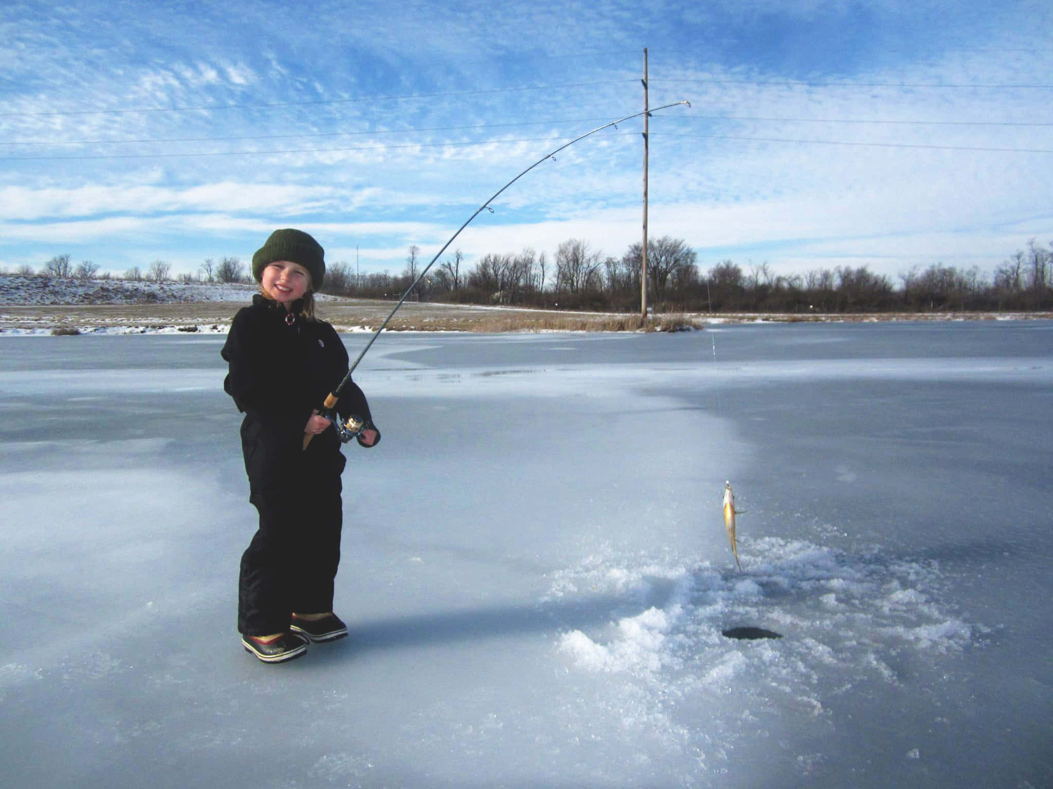 Ice Fishing 桌布 壁紙 — 一個微笑的孩子穿著冬季裝備站在結冰的湖上，手持釣魚竿，一條釣到的魚從冰洞中垂下來，在明亮的藍天下。