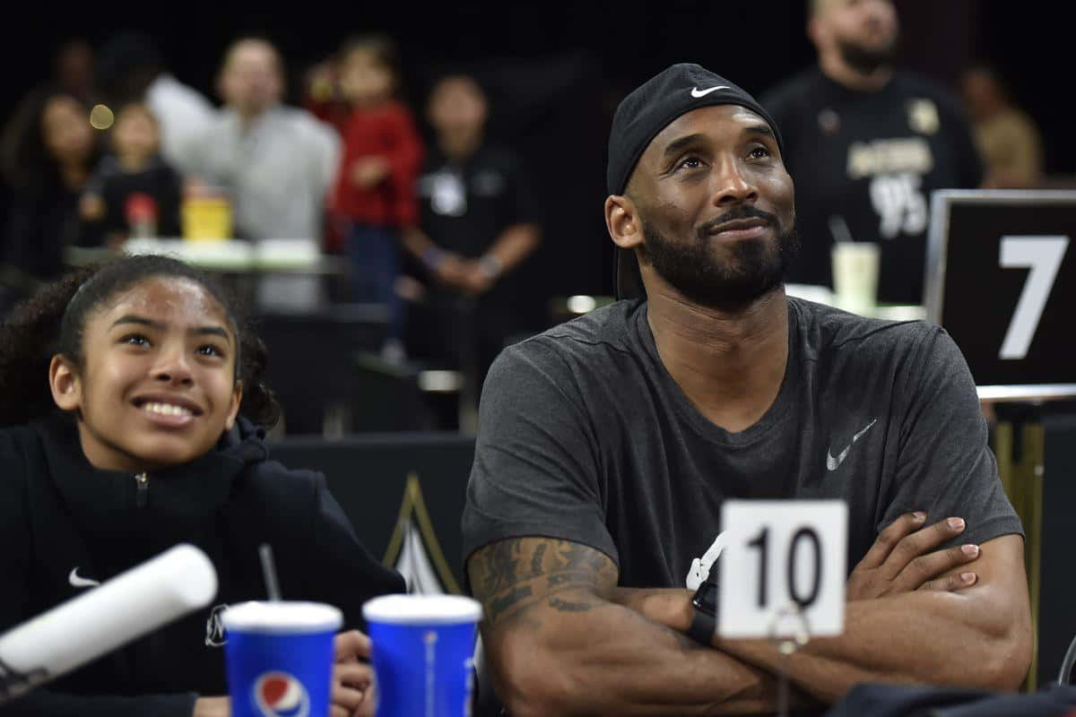 Kobe And Gigi 桌布 壁紙 — 一個微笑的年輕女孩和一個戴黑色鴨舌帽的鬍鬚男人一起坐在籃球比賽場上，背後是模糊的觀眾。