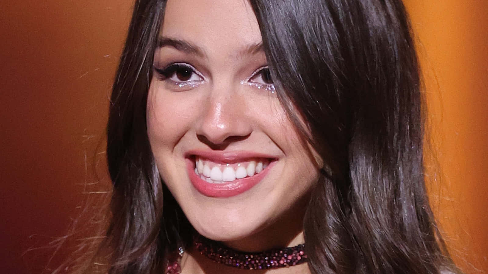 Olivia Rodrigo 桌布 壁紙 — 一個微笑的年輕女性特寫肖像，深色頭髮，閃亮眼妝，背景是溫暖的橙色。
