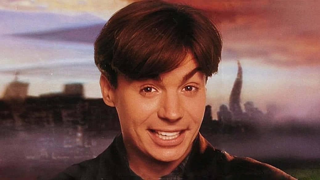 Mike Myers 桌布 壁紙 — 一個微笑的棕髮男子，背景是日落時分模糊的城市天際線，帶有溫暖的橙色和紫色色調。