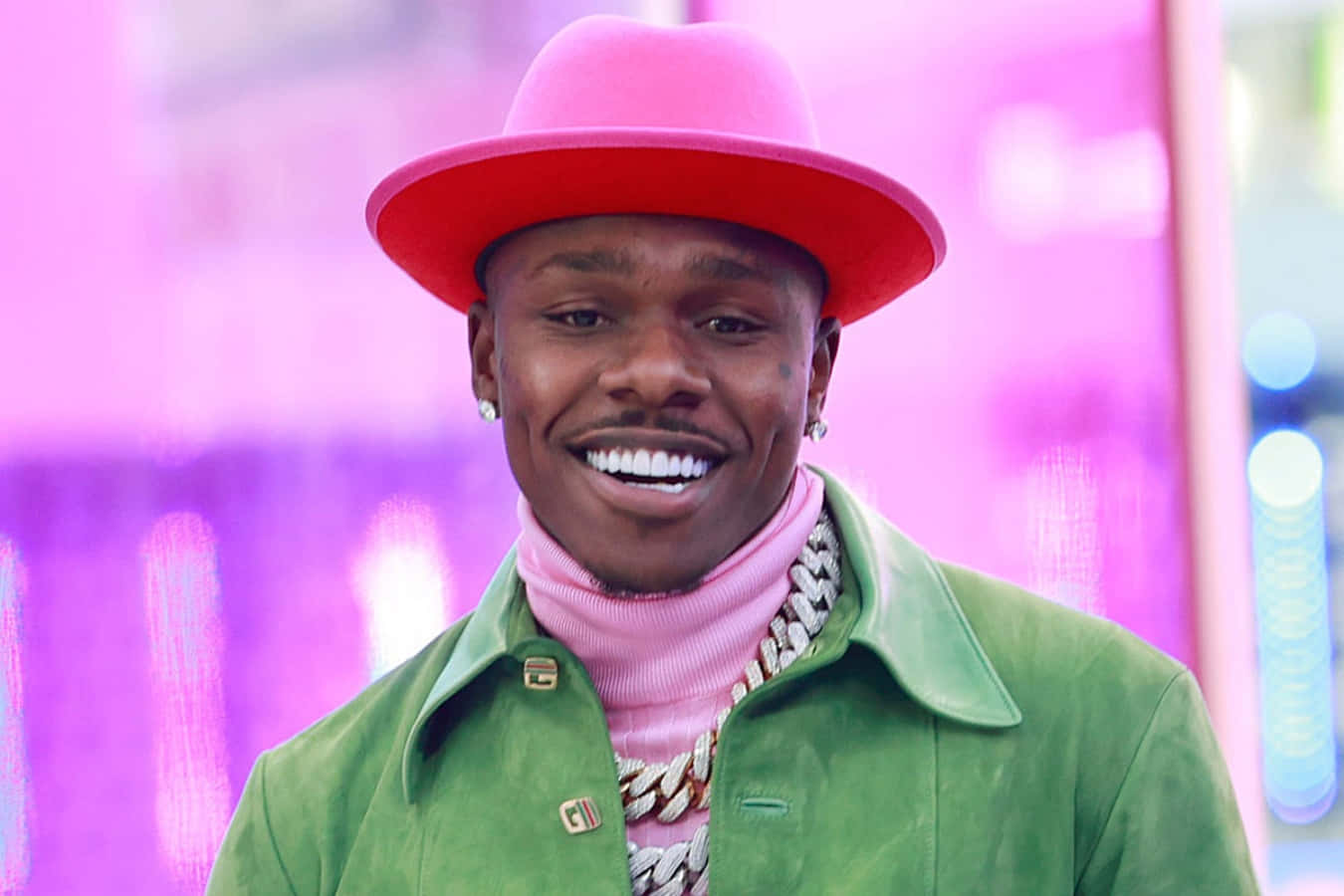 Dababy 桌布 壁紙 — 一個微笑的男人，戴著亮粉色帽子、綠色夾克和粉色高領毛衣，站在發光的粉紫色城市背景前。
