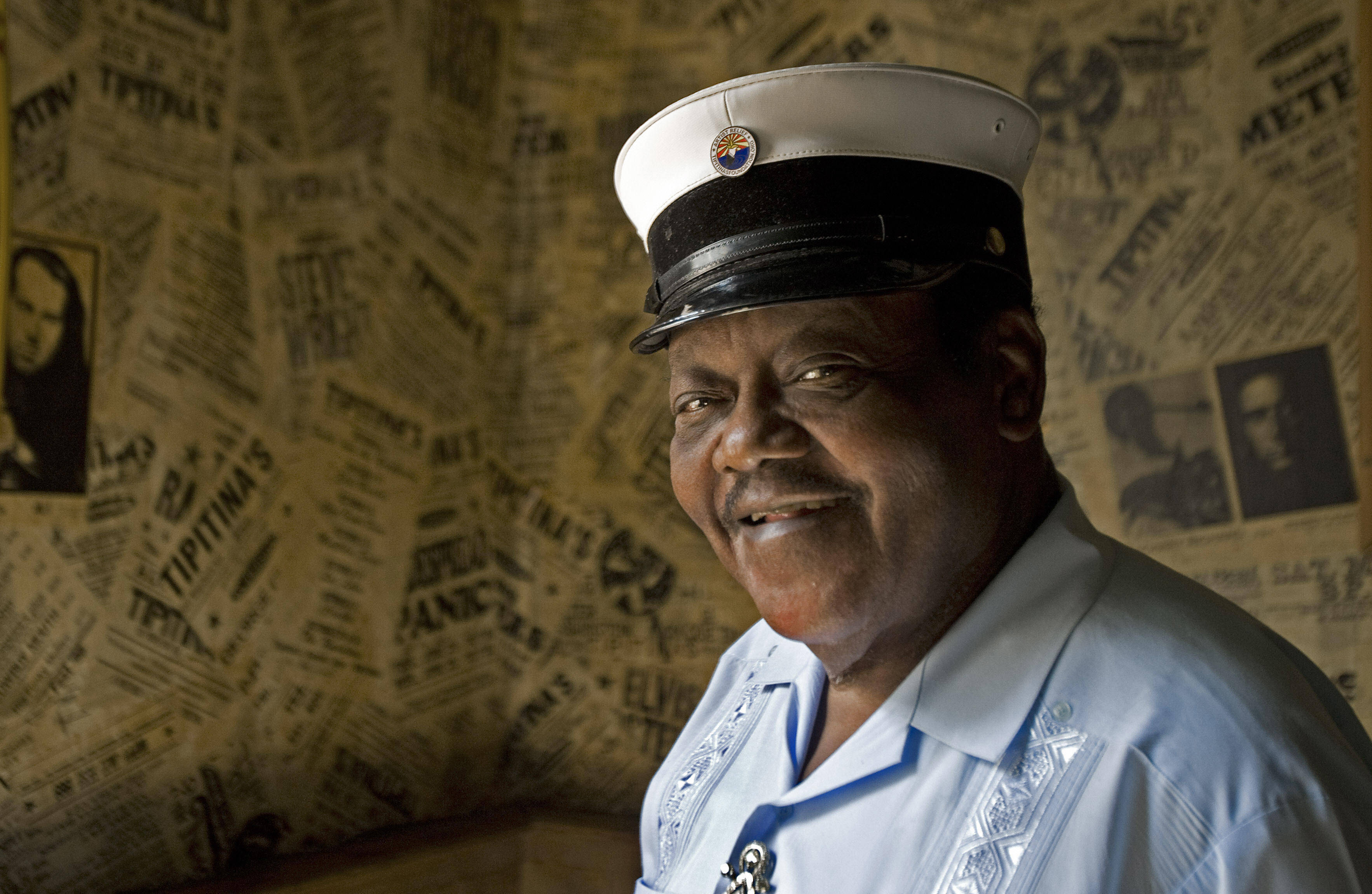 Fats Domino Antoine Dominique Domino Jr 壁紙 — 一個微笑的男人，戴著白色水手帽和淺藍色襯衫，站在一面貼滿復古報紙剪報的牆前。