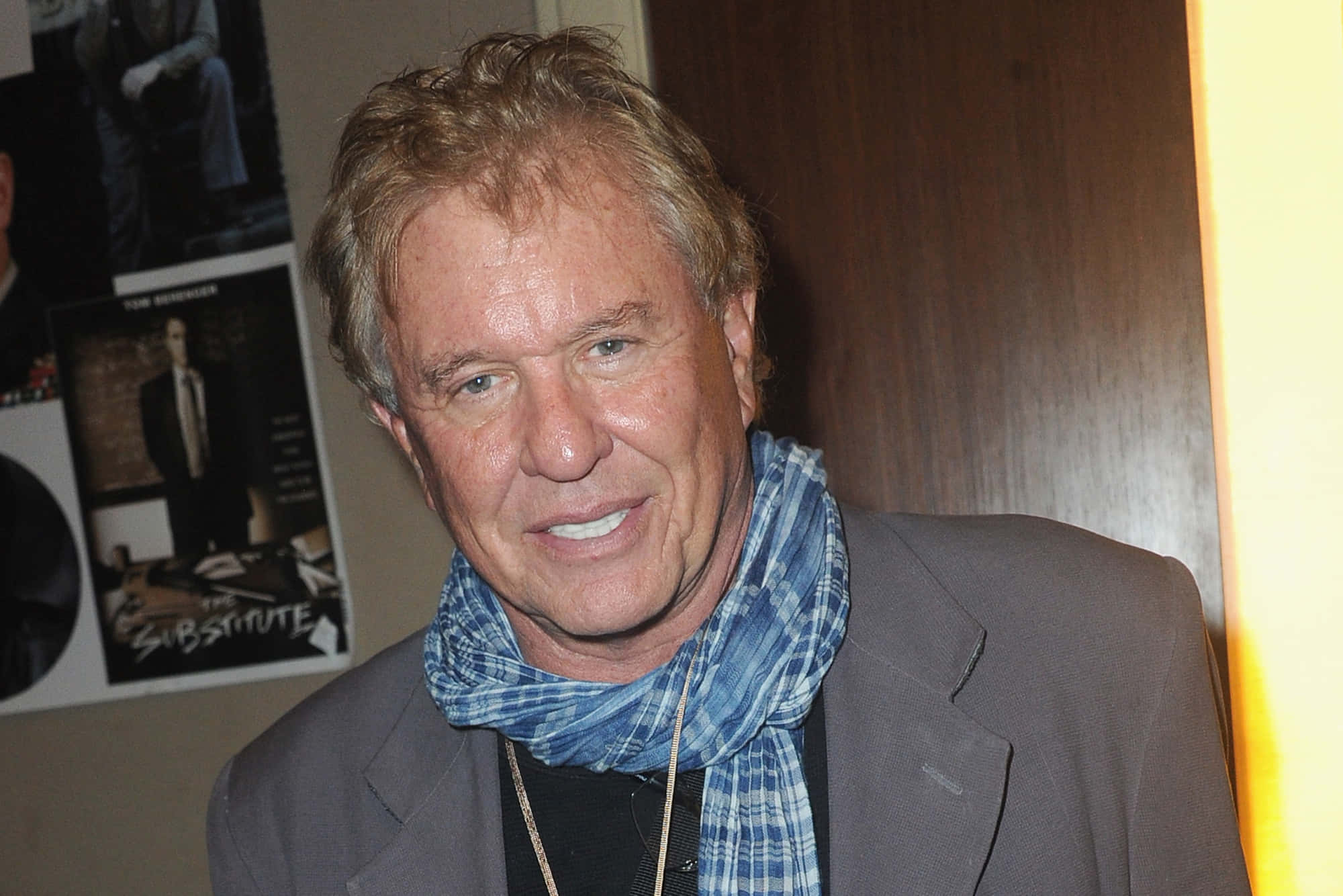 Tom Berenger 桌布 壁紙 — 一個微笑的男人，波浪金髮，戴著藍色格紋圍巾和灰色西裝外套，背景是海報。